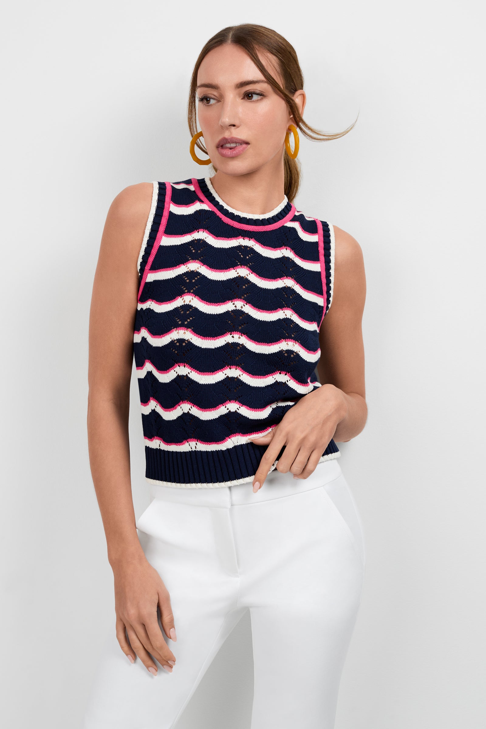 A woman models the Cawley Knit Tank—a sleeveless navy blue sweater with white and pink wavy stripes—paired with white high-waisted pants and large yellow hoop earrings against a plain white background.