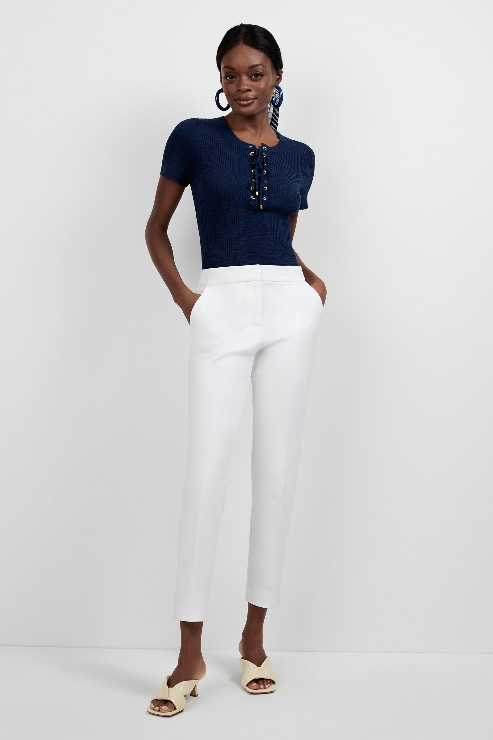 A woman stands against a white background, wearing a short-sleeve navy ribbed top with lace-up front, blue hoop earrings, cream heeled sandals, and the Aubree 2 Cigarette Pants in white. Her hands are in her pockets.