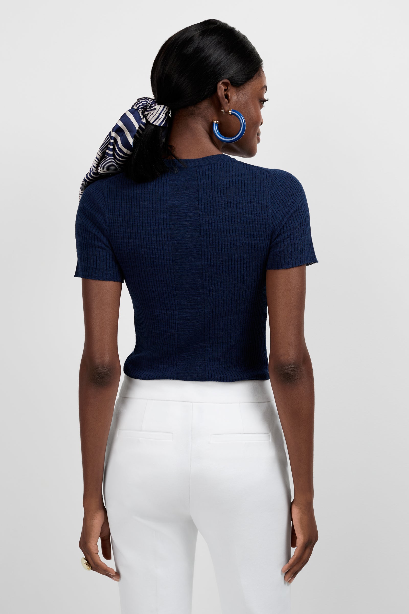 A woman faces away, wearing the Mina Lace-Up Top in navy, white high-waisted pants, blue hoop earrings, and a striped hair scarf tied around her ponytail.