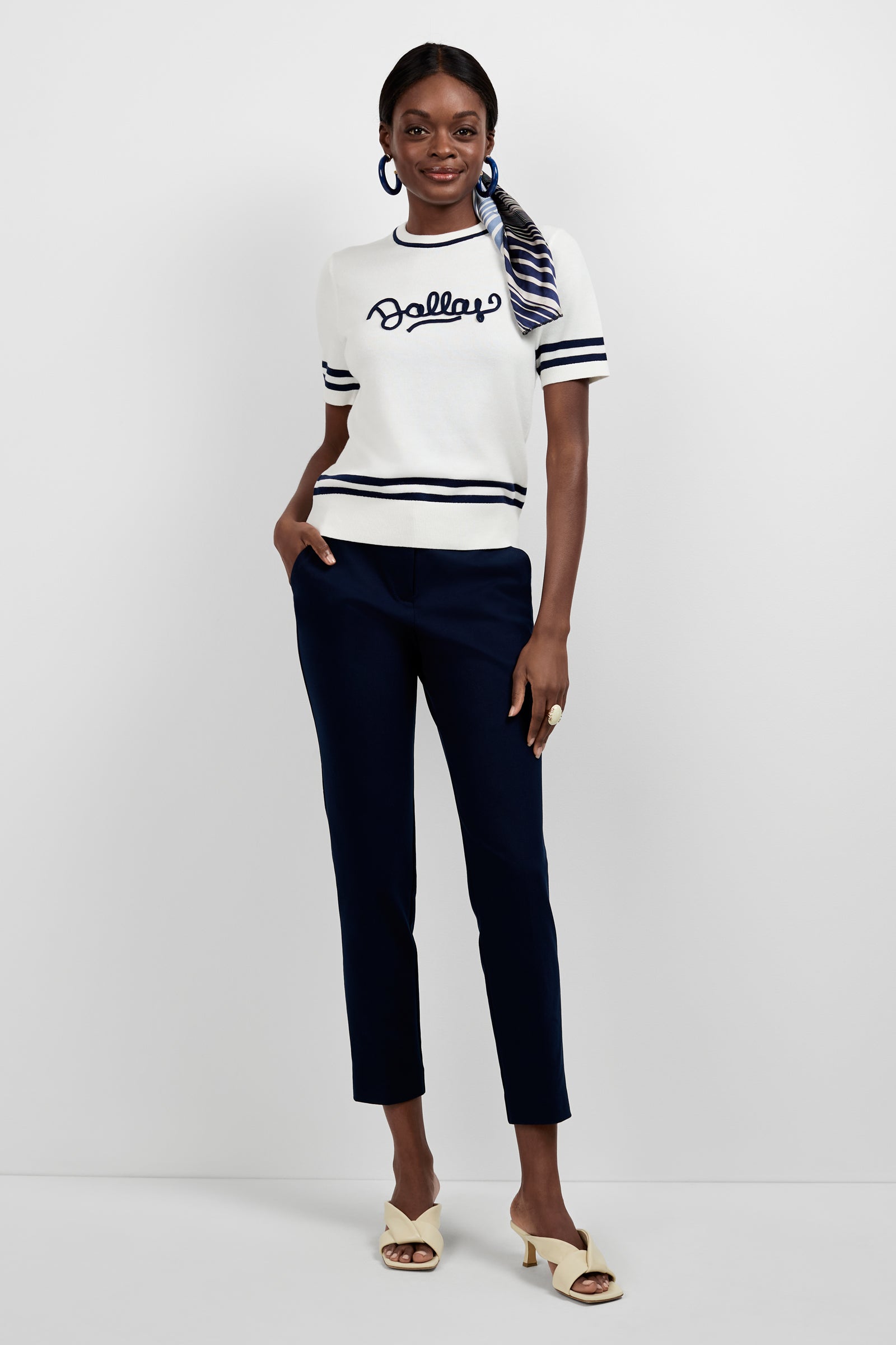 A woman poses against a white background wearing a white “Dallas” short-sleeve top, Aubree 2 Cigarette Pants in navy blue, cream heeled sandals, hoop earrings, and a striped scarf tied in her hair.