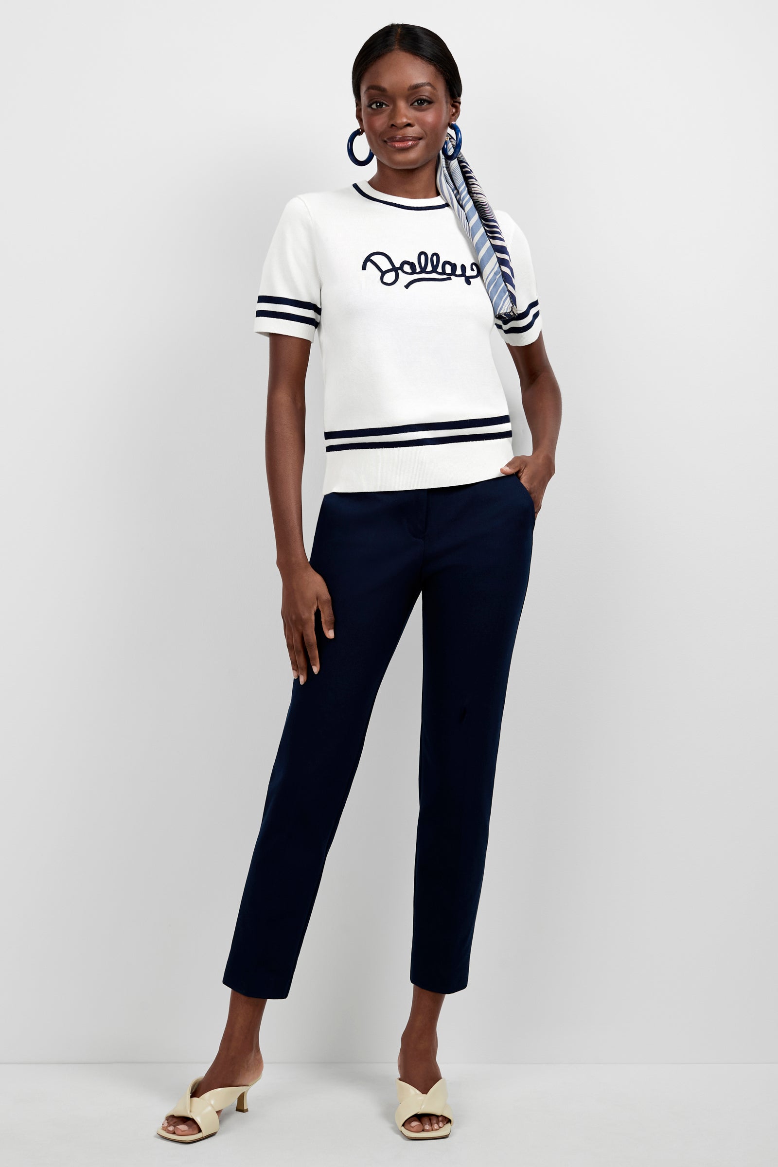A woman stands against a white background wearing the Old Fashioned Sweater -Dallas, navy blue pants, white heeled sandals, hoop earrings, and a striped scarf tied in her hair.