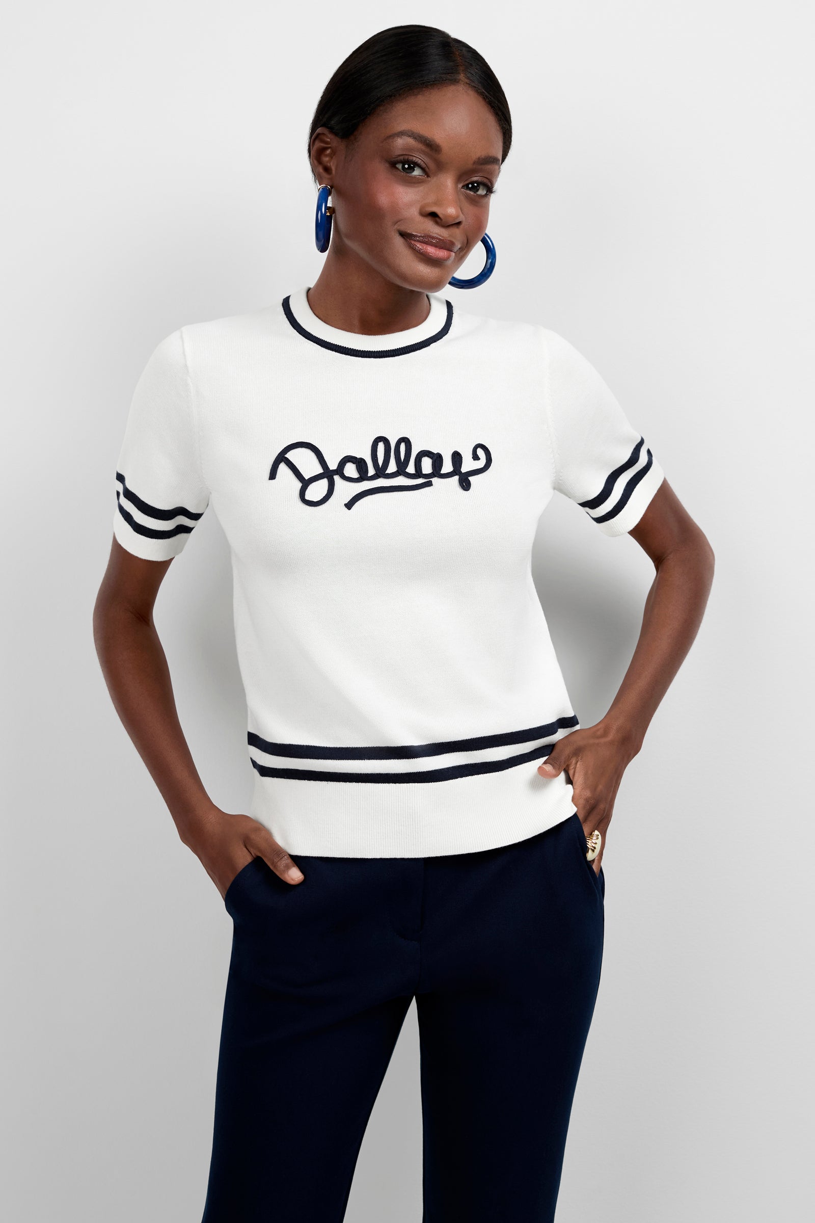 A woman poses confidently against a plain light background wearing the Old Fashioned Sweater - Dallas, a white short-sleeve sweater with black trim and "Jolly" across the chest, paired with dark navy pants.