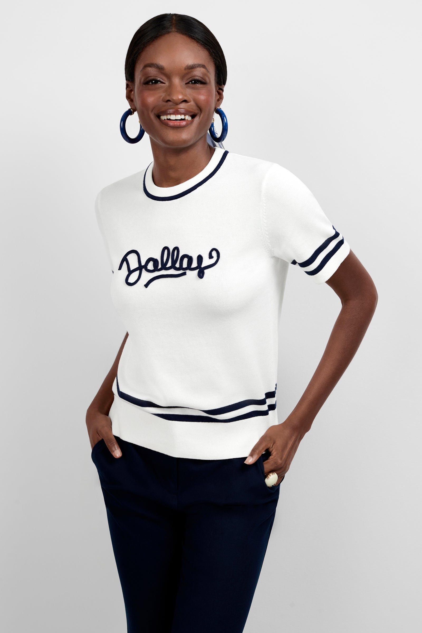 A smiling woman wears the Old Fashioned Sweater -Dallas, a white short-sleeve sweater with navy trim and "Dallas" on the front, paired with navy pants and large blue hoop earrings, posing with her hands in her pockets.