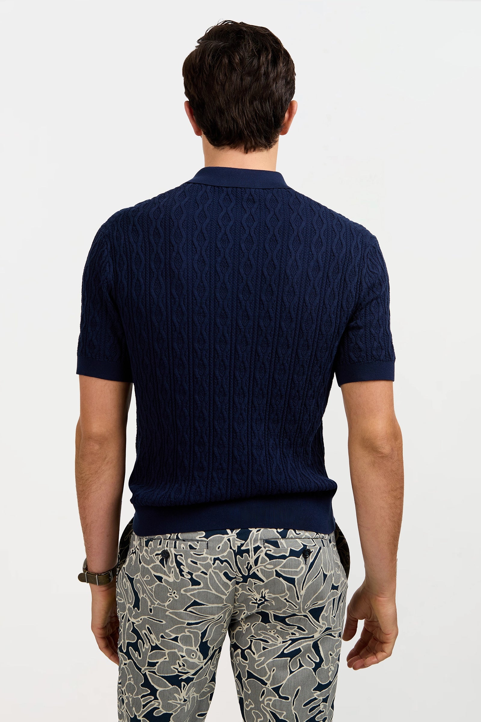 A man stands with his back to the camera, wearing the Elio Short-Sleeve Polo in navy blue and light-colored pants with a bold dark floral pattern against a plain white background.