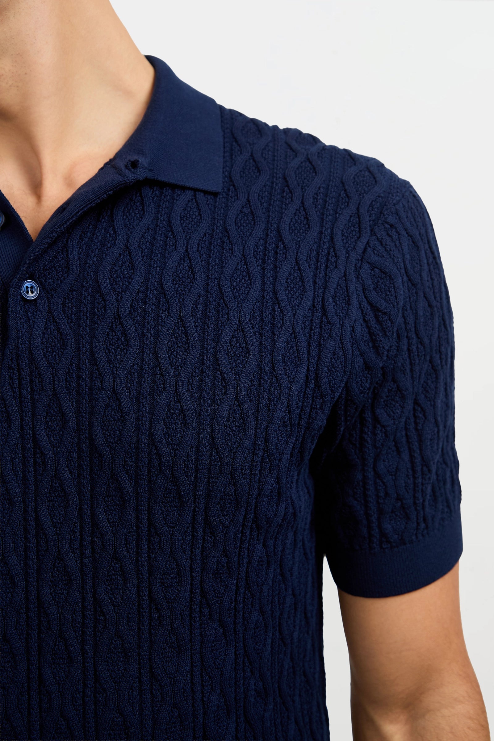 A close-up of a person wearing the Elio Short-Sleeve Polo in navy blue, featuring a collar, buttons, and cable knit texture, highlighting the shirt’s pattern along with part of the neck and arm.