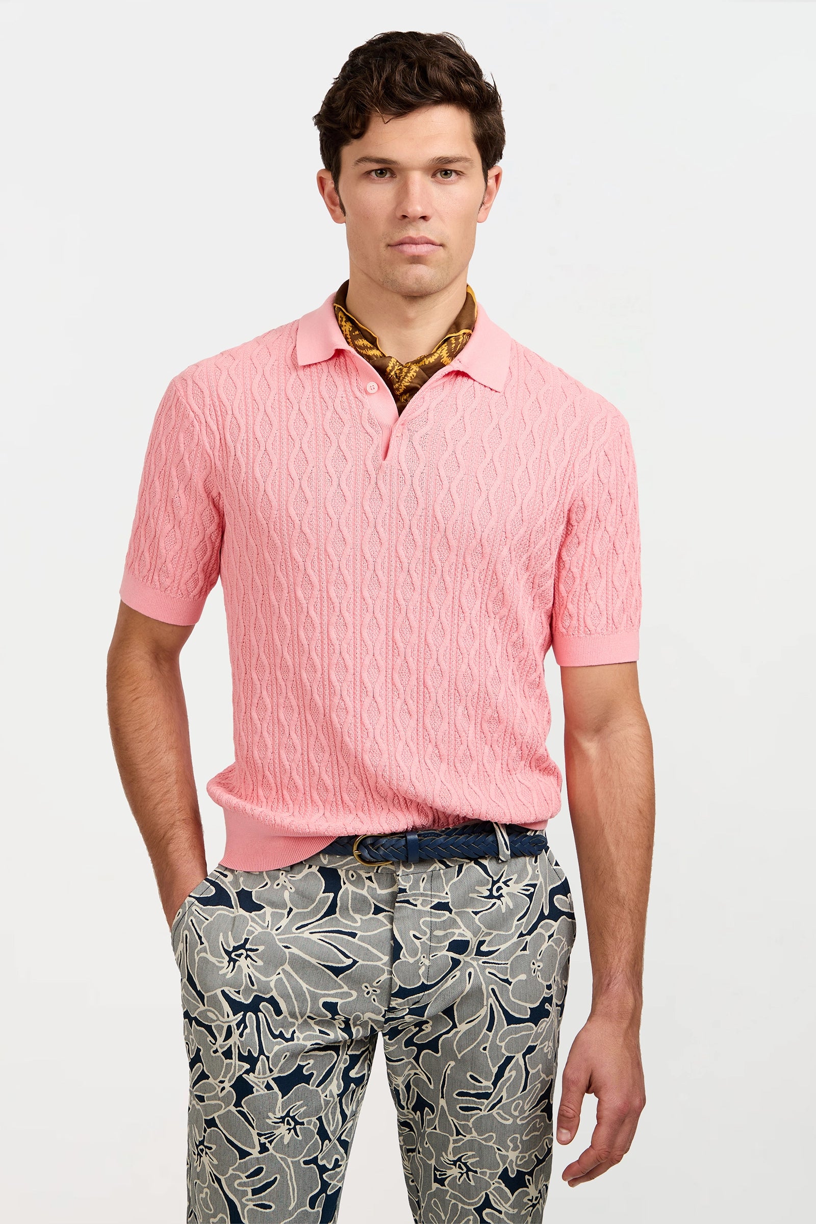 A young man stands with hands in his pockets against a plain background, wearing the Elio Short-Sleeve Polo in light pink, patterned blue and white floral pants, and a gold scarf around his neck.