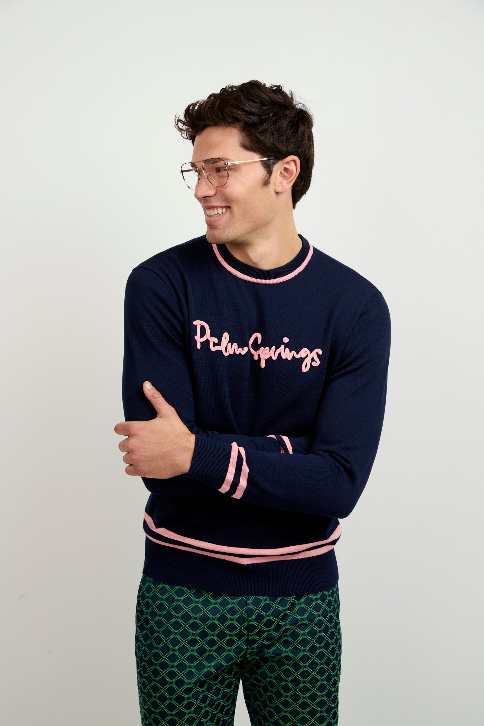 A man with short brown hair and glasses smiles while looking to the side. He wears the Palm Springs Crewneck Sweater in navy blue, paired with green patterned pants, standing against a plain light background.