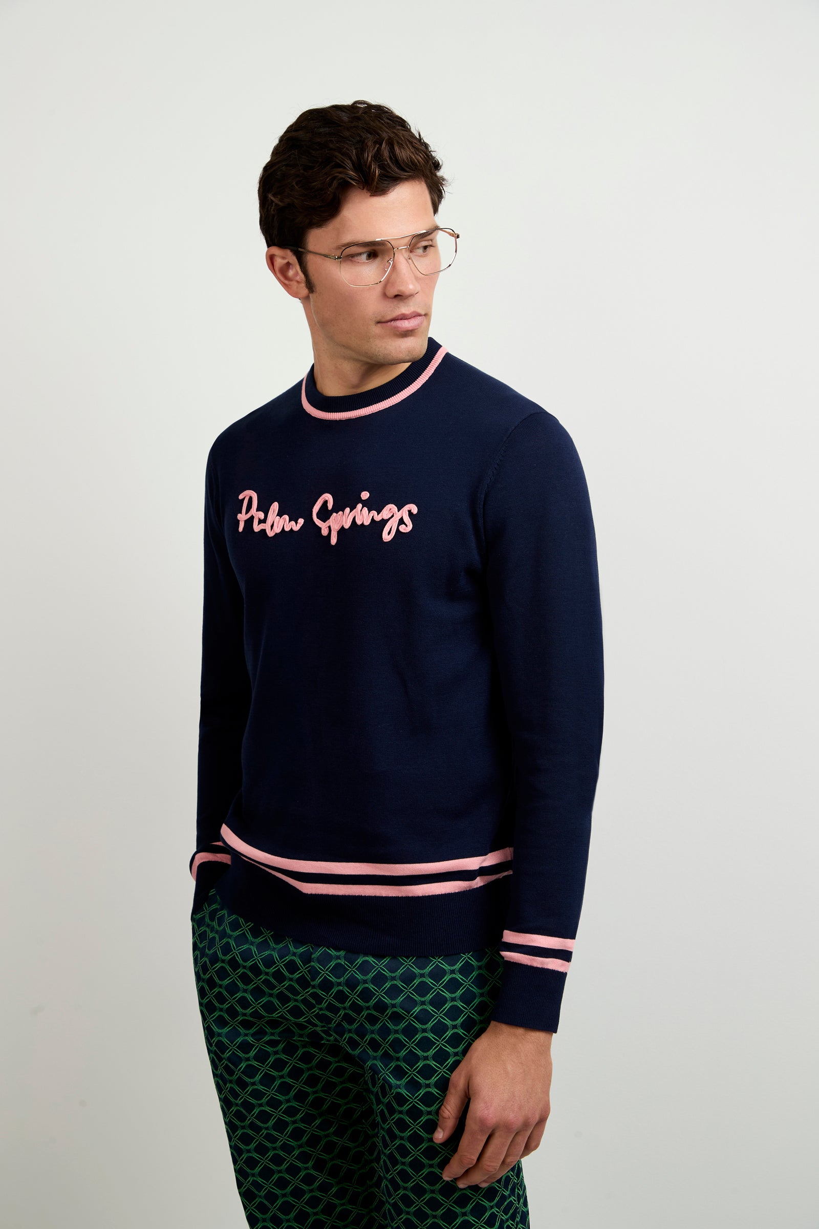 A man in glasses and a navy blue Palm Springs Crewneck Sweater with pink trim and green patterned pants stands against a light background, looking to the side.