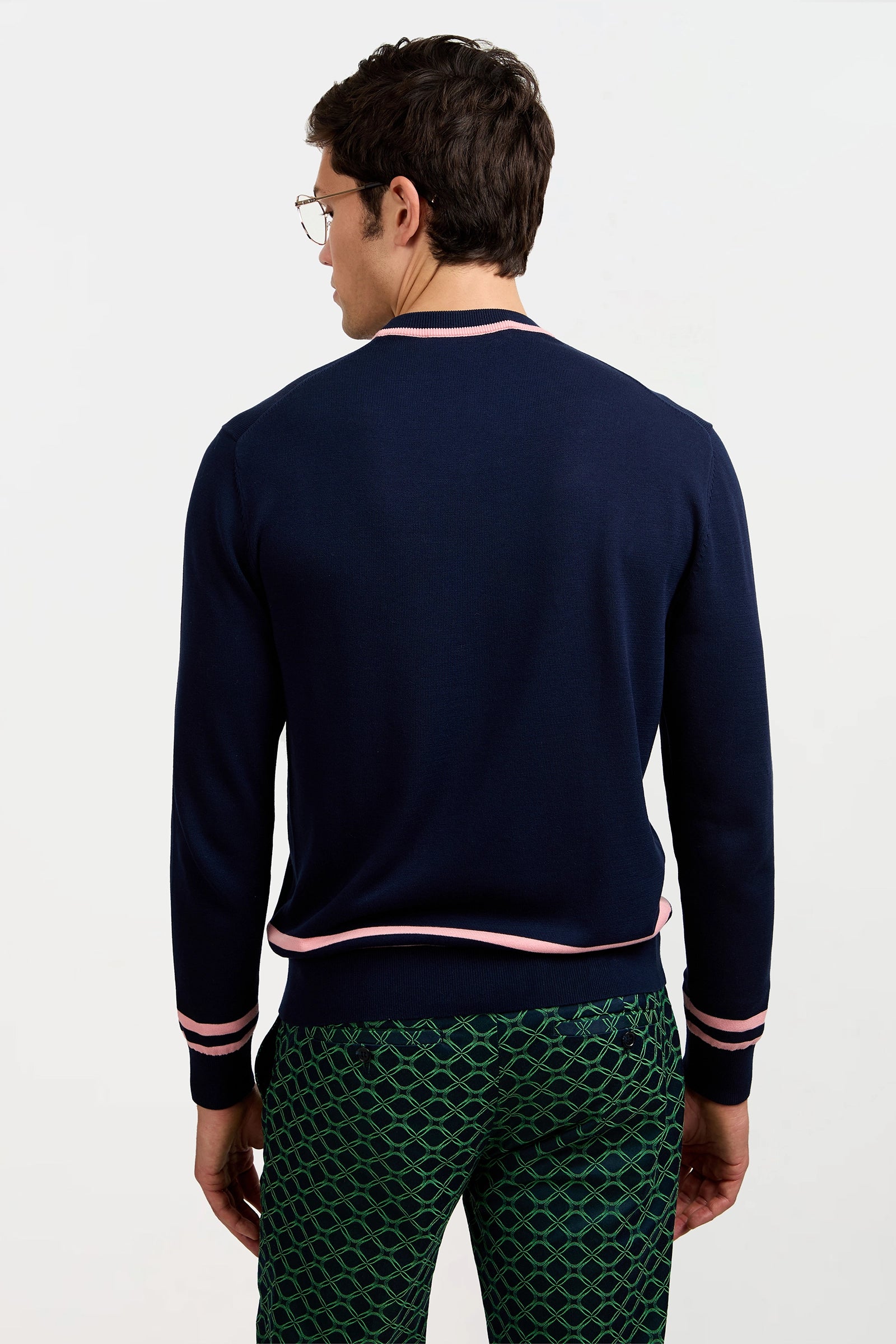 A man with short brown hair and glasses, wearing a Palm Springs Crewneck Sweater in navy with pink trim and green patterned pants, stands facing away from the camera against a plain white background.