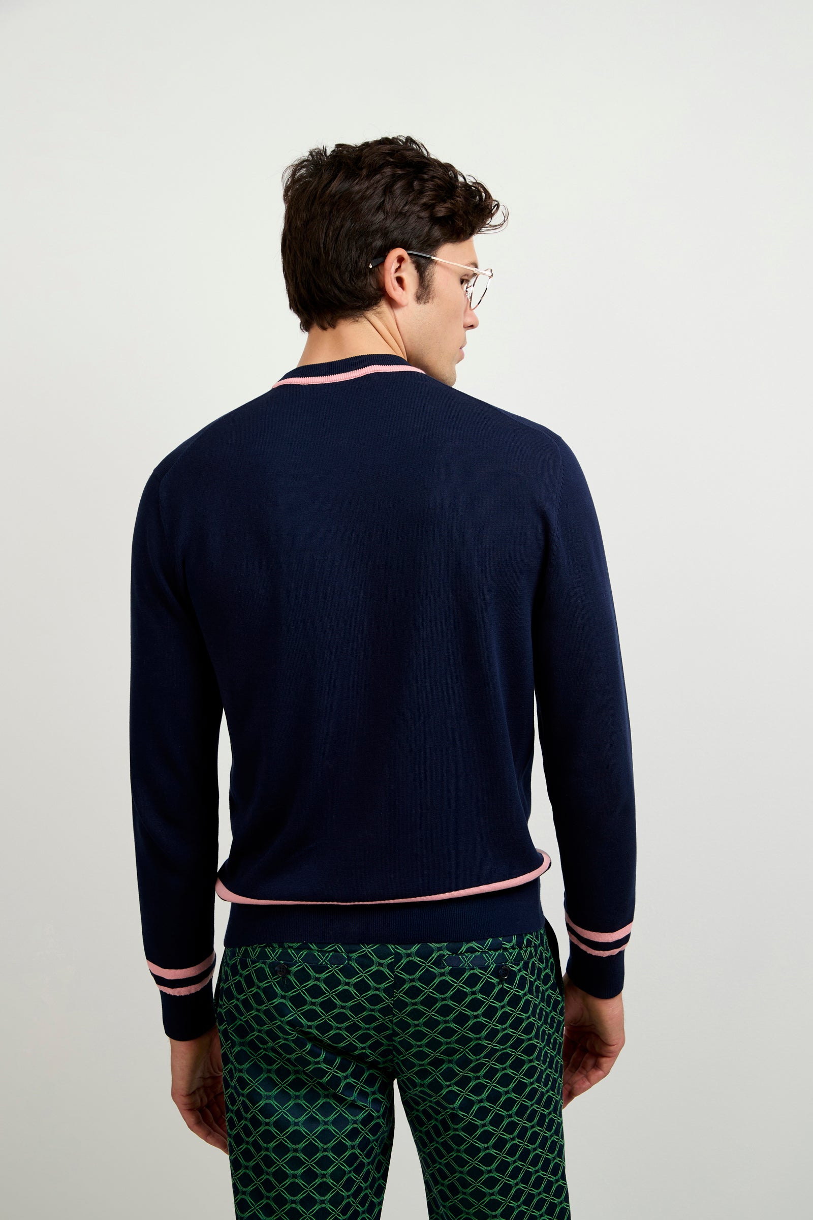 A man with short brown hair and glasses, wearing the Palm Springs Crewneck Sweater in navy blue with pink trim and patterned green pants, stands facing away from the camera against a plain light background.