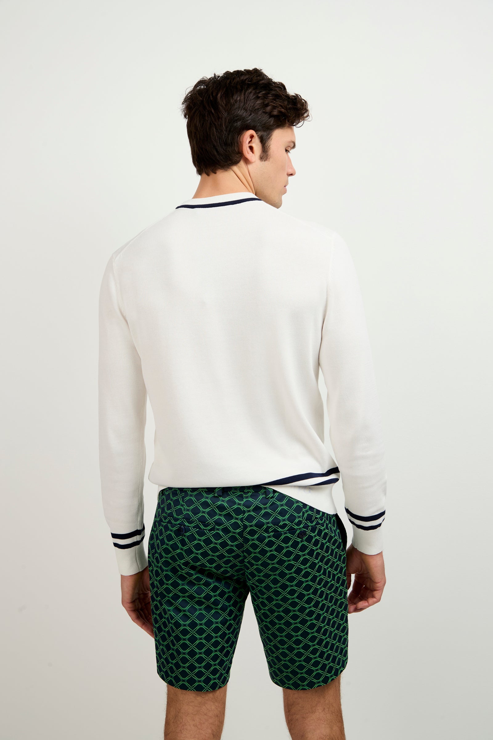 A man faces away from the camera wearing the Palm Springs Crewneck Sweater, a white long-sleeve with dark trim, paired with green patterned shorts. The background is plain and light-colored.