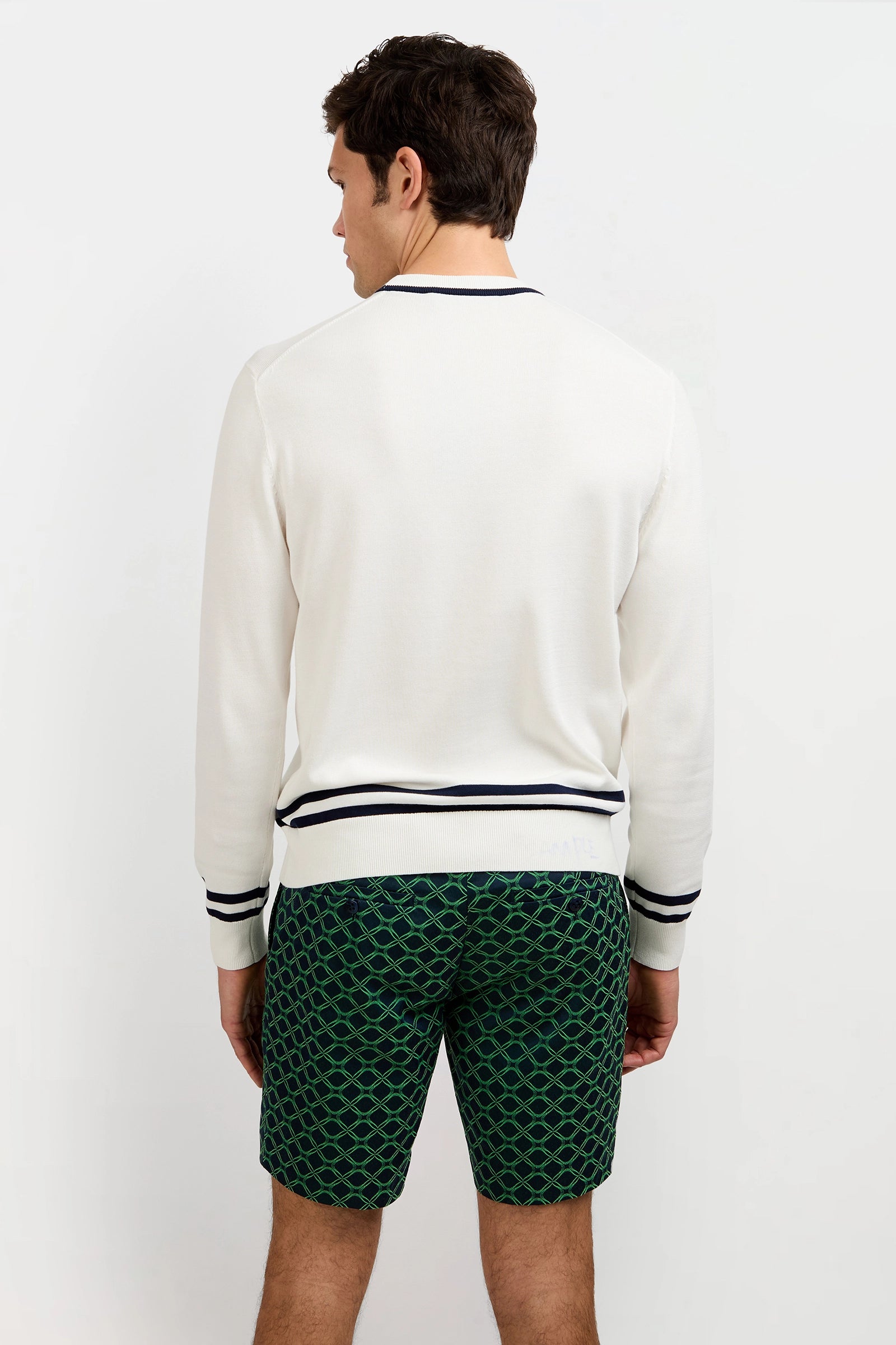 A man stands facing away, wearing the Palm Springs Crewneck Sweater with navy trim and green patterned shorts. The background is plain white.
