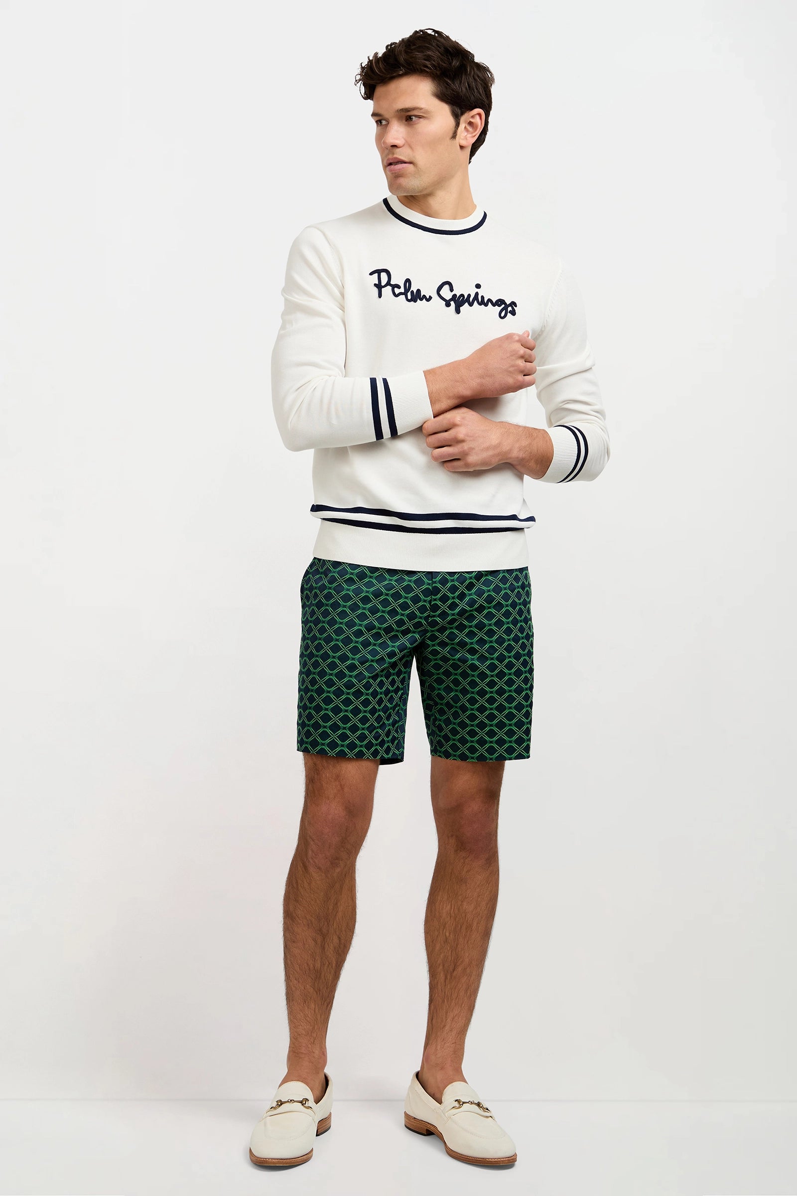 A man with short dark hair stands against a plain background wearing green Lawrence Short - Clover Geo Jacquard (Ink/Multi) patterned shorts, a white Palm Springs sweatshirt, and white loafers, looking to the side with arms slightly crossed.