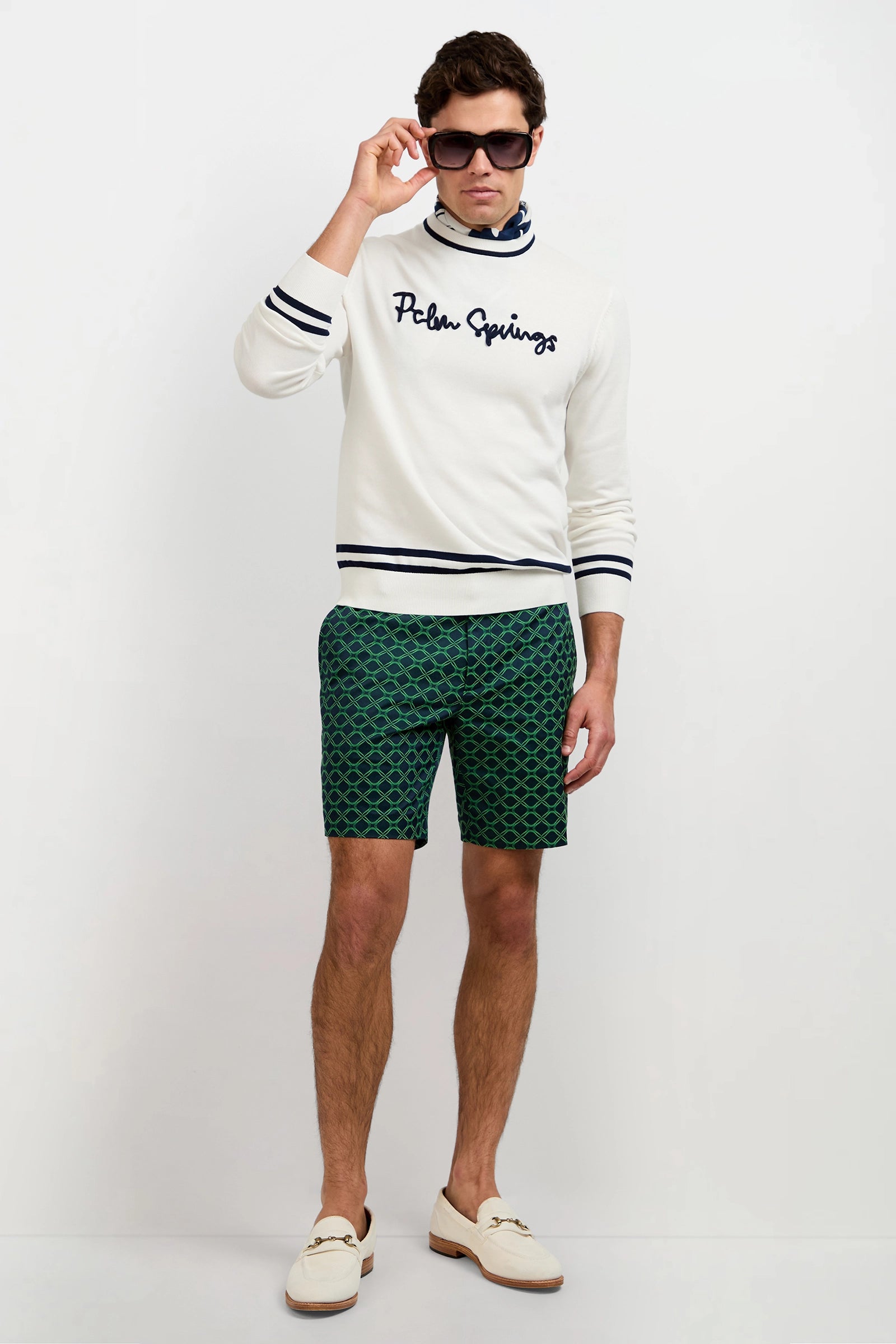 A man stands against a white background wearing sunglasses, green patterned shorts, cream loafers, and the Palm Springs Crewneck Sweater. He lifts his sunglasses slightly with one hand.