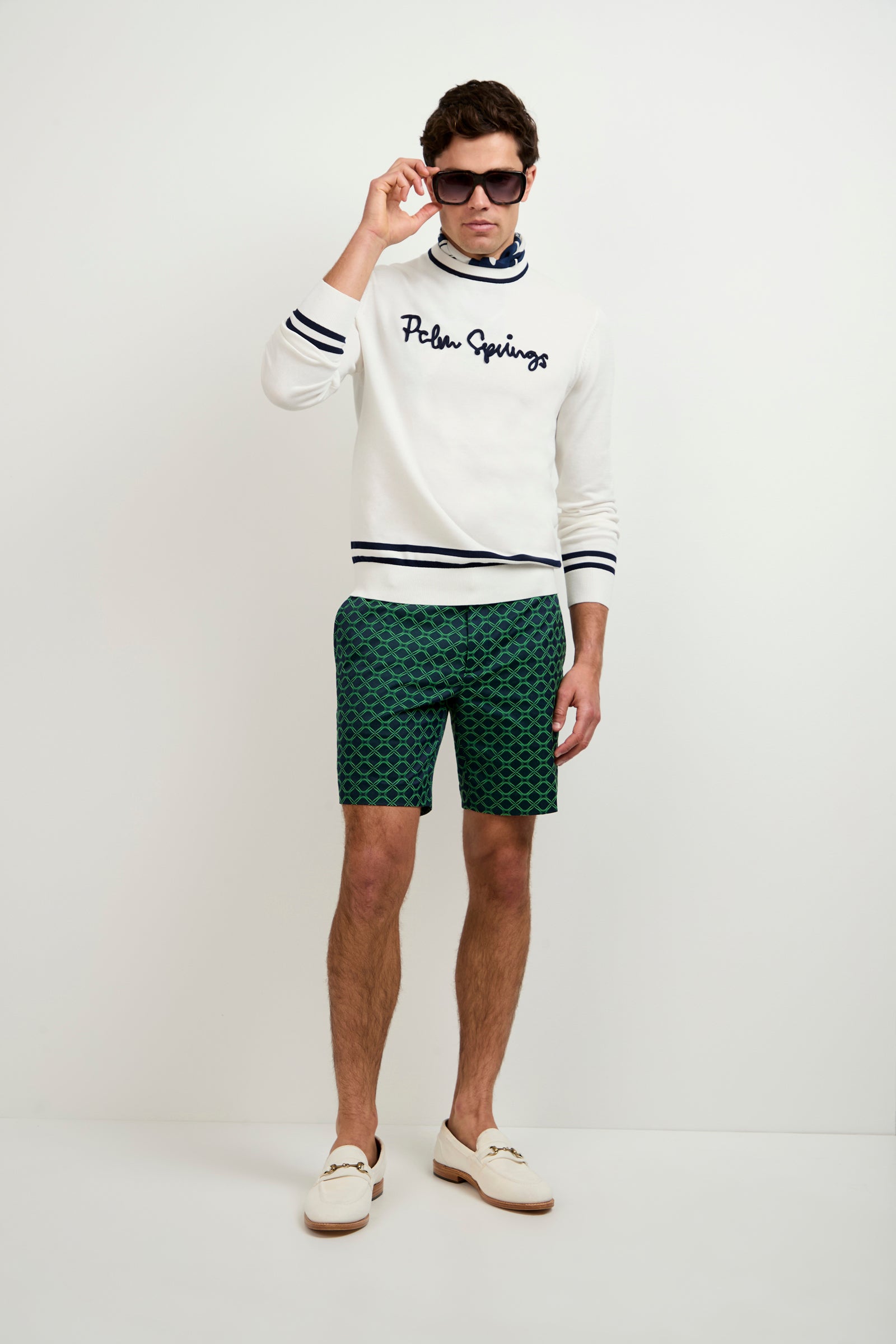 A man stands against a white background wearing dark sunglasses, a white Palm Springs sweater, Lawrence Short - Clover Geo Jacquard green patterned shorts, and white loafers. He holds his sunglasses with one hand.