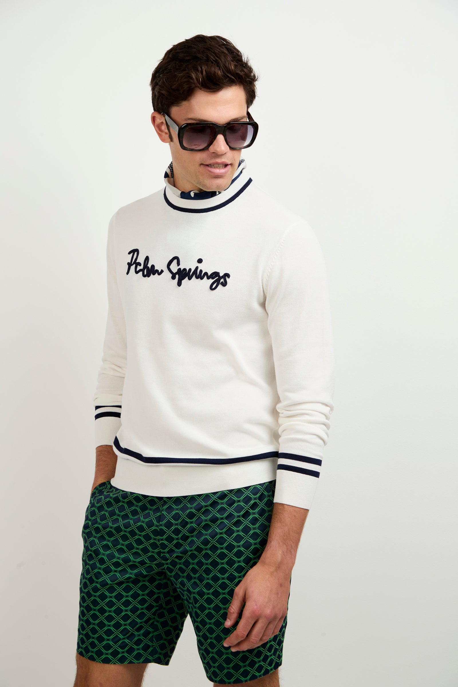 A man stands against a plain white background wearing large sunglasses, green patterned shorts, and the Palm Springs Crewneck Sweater.