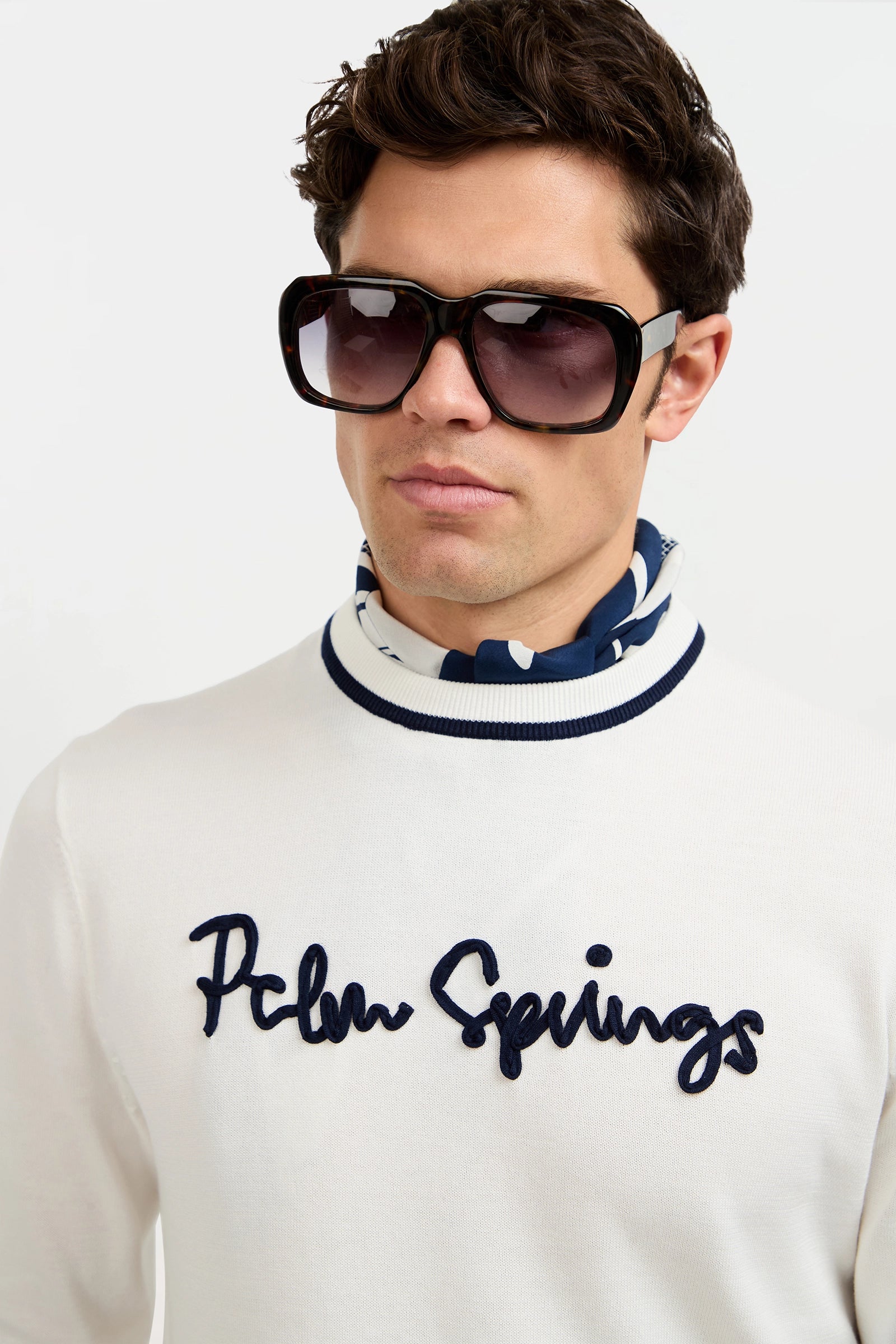 A man stands against a plain white background wearing oversized dark sunglasses, a blue and white patterned scarf, and the Palm Springs Crewneck Sweater.
