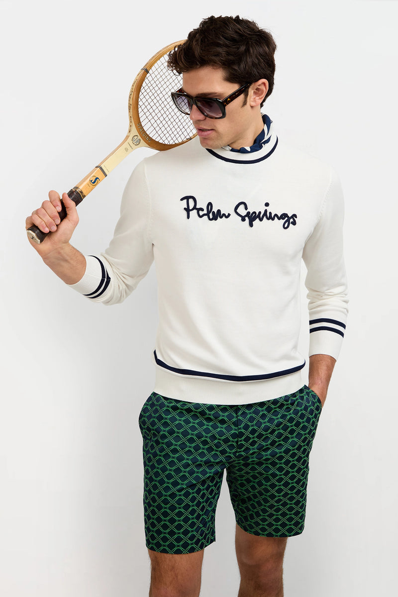 A man wearing dark sunglasses, green patterned shorts, and the Palm Springs Crewneck Sweater poses with a vintage tennis racket over his shoulder against a plain white background.