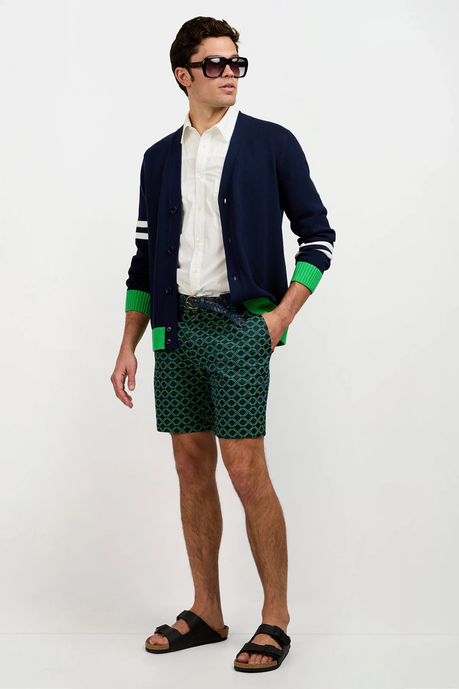 A man wears dark sunglasses, a white shirt, the Amadeo Cardigan with green cuffs, patterned green shorts, and black sandals as he stands with one hand in his pocket against a plain white background.