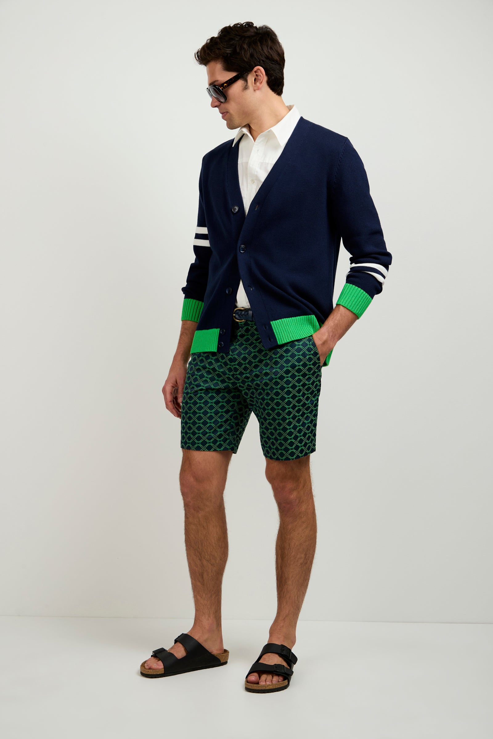 A man wearing sunglasses, the Amadeo Cardigan with green and white accents over a white shirt, green patterned shorts, and black sandals stands against a plain white background.