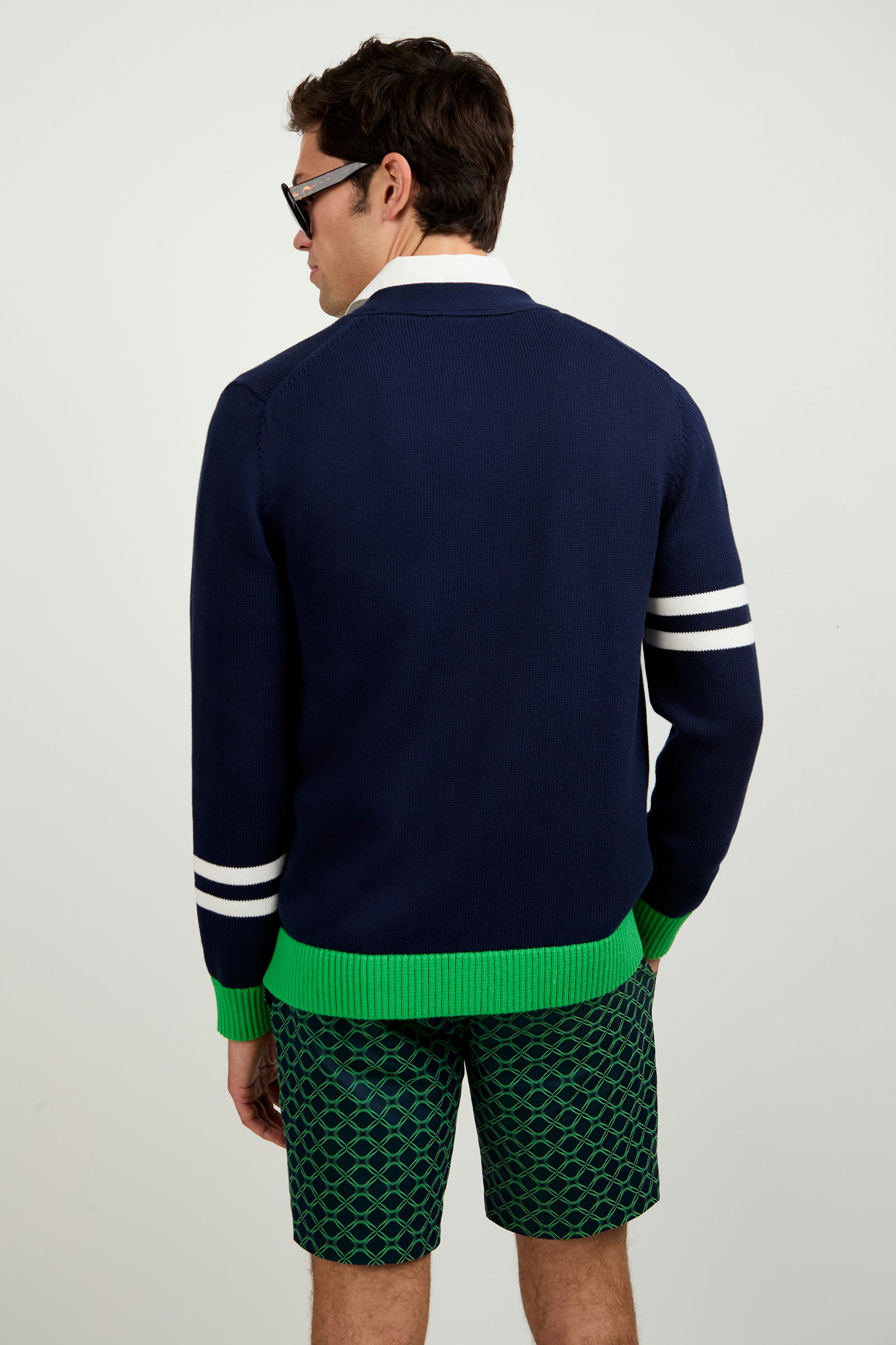 A man with dark hair and sunglasses faces away, wearing the Amadeo Cardigan—navy blue with two white sleeve stripes and a green hem—paired with green patterned shorts, set against a plain white background.