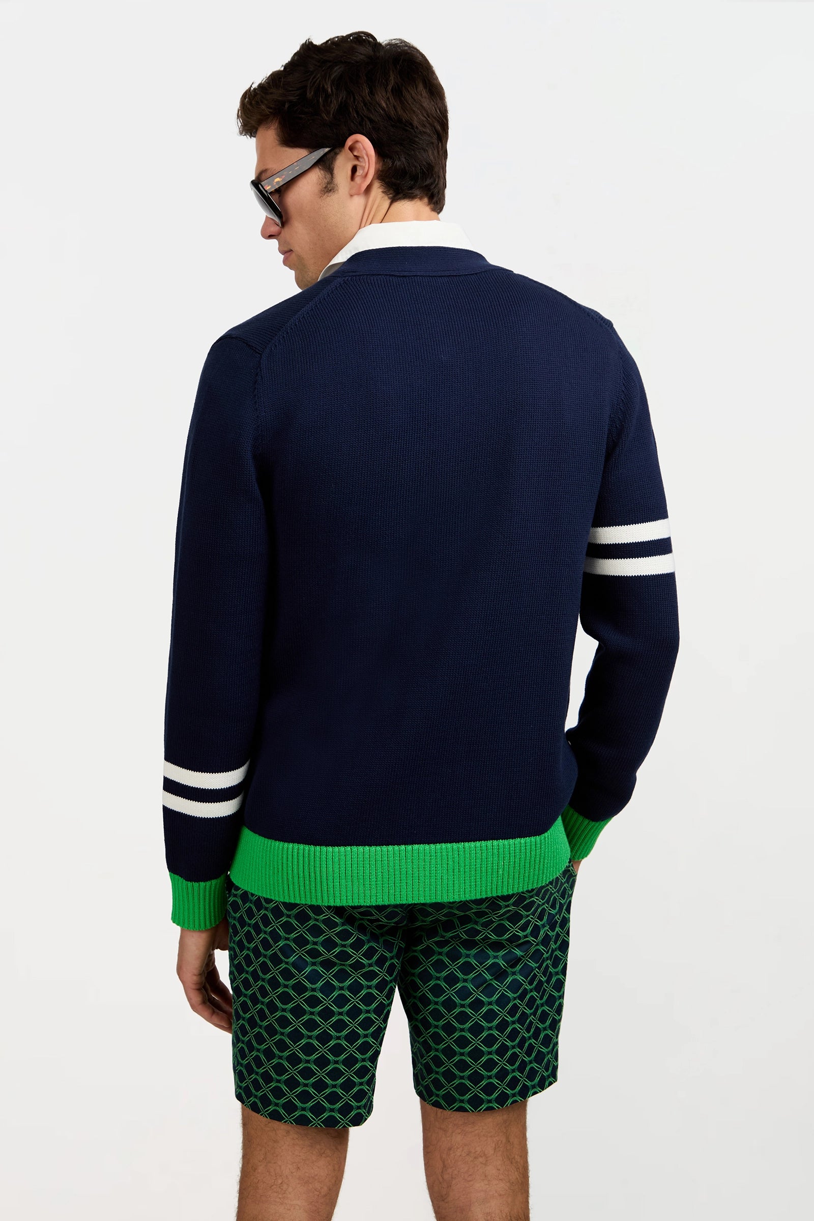 A man stands facing away, wearing the Amadeo Cardigan—navy with white sleeve stripes, a white collar, and green trim—paired with green patterned shorts and glasses against a plain white background.