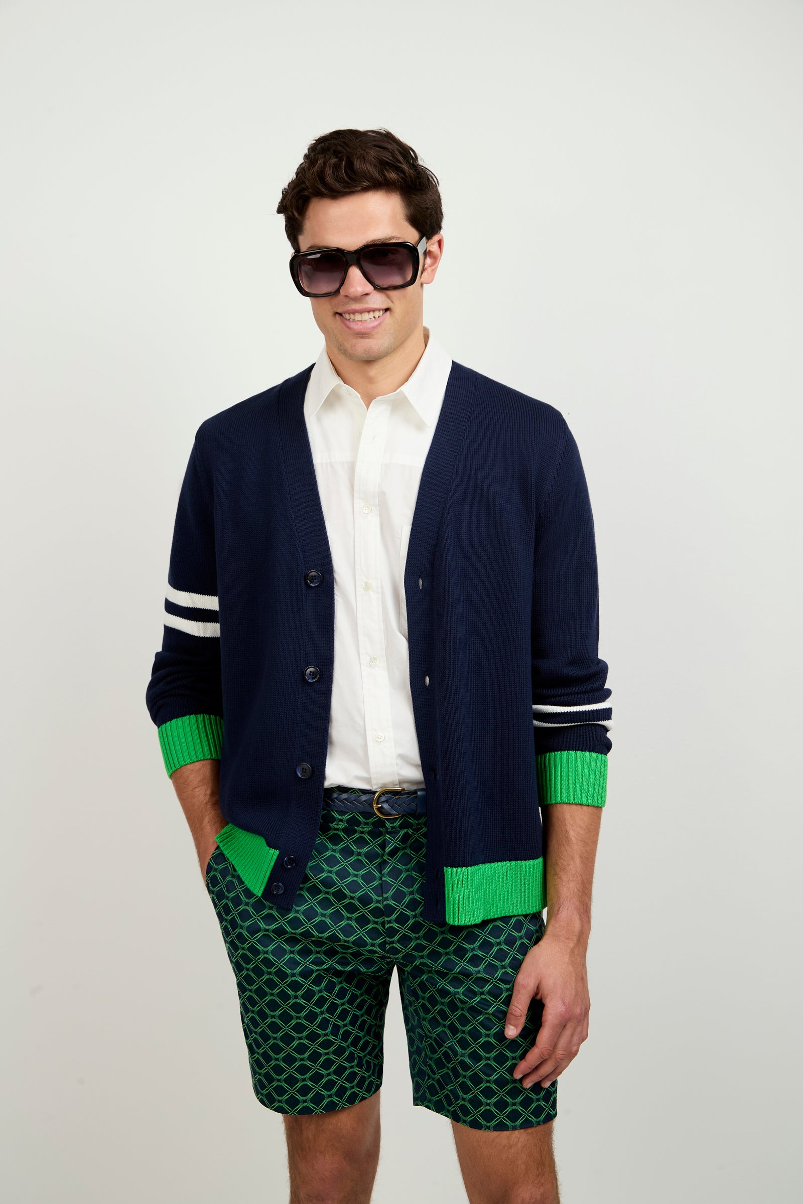 A man smiles at the camera with hands in his pockets, wearing large black sunglasses, a white shirt, patterned green shorts, and the Amadeo Cardigan in INK MULTI.