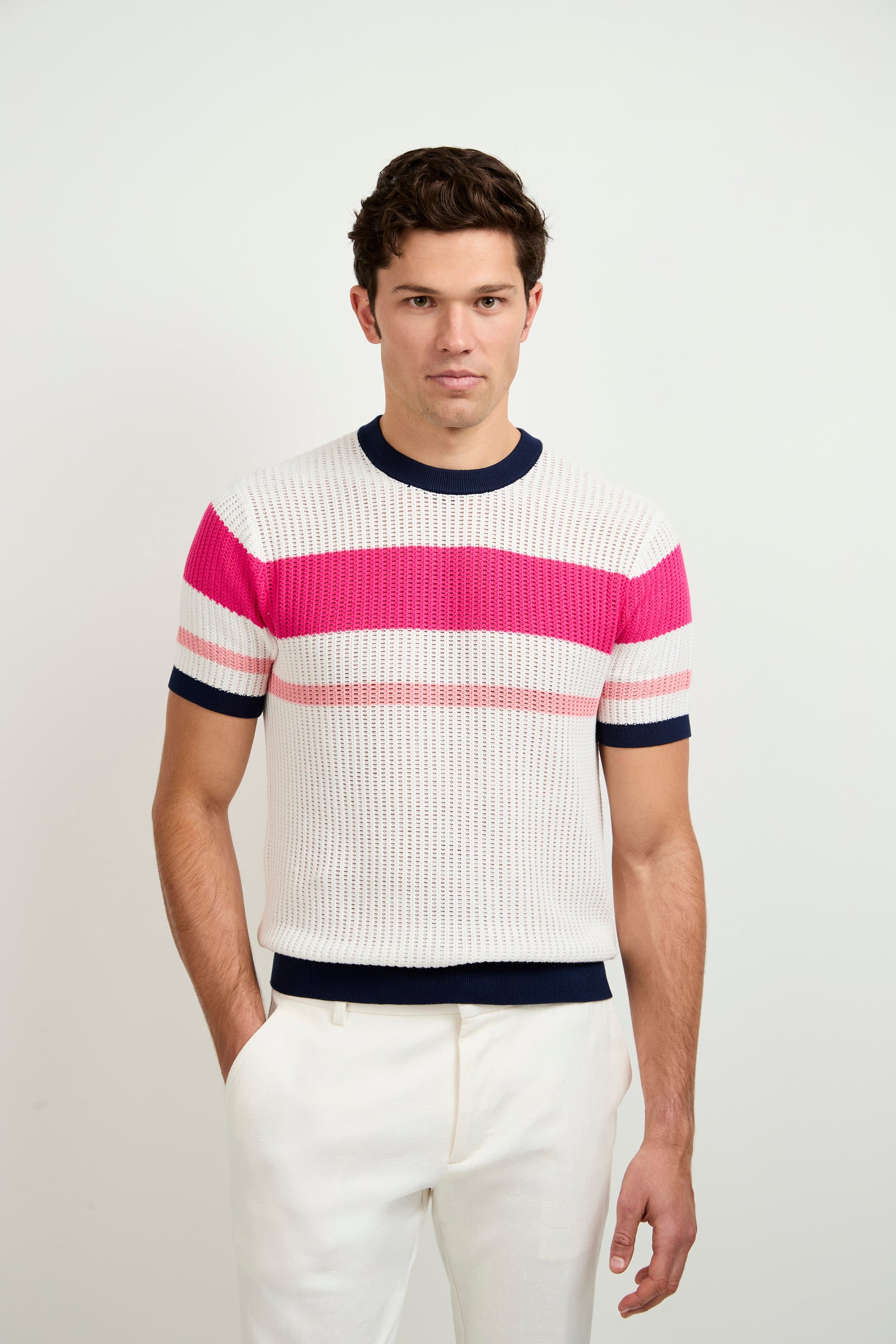 A man with short brown hair and a neutral expression stands against a white background wearing the Aldo Short-Sleeve Henley, which features pink, white, and navy stripes, paired with white pants.