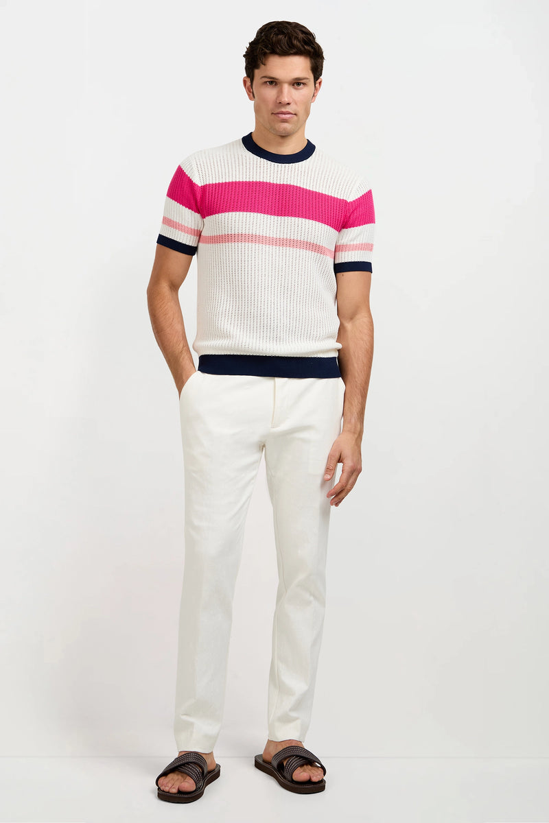 A man stands against a white background wearing the Aldo Short-Sleeve Crewneck—white with pink and red horizontal stripes—paired with white pants and dark sandals.
