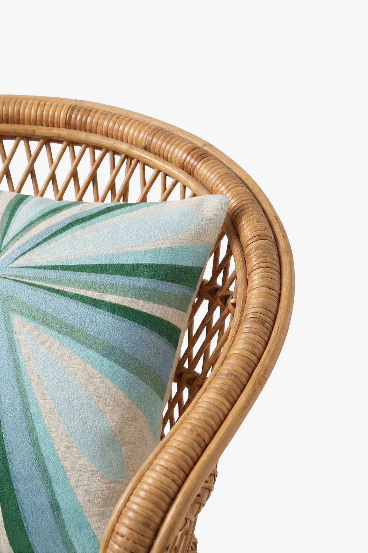 A close-up of a wicker chair with the Shante Embroidered Square Pillow, featuring green, blue, and cream abstract petal shapes on a white background.