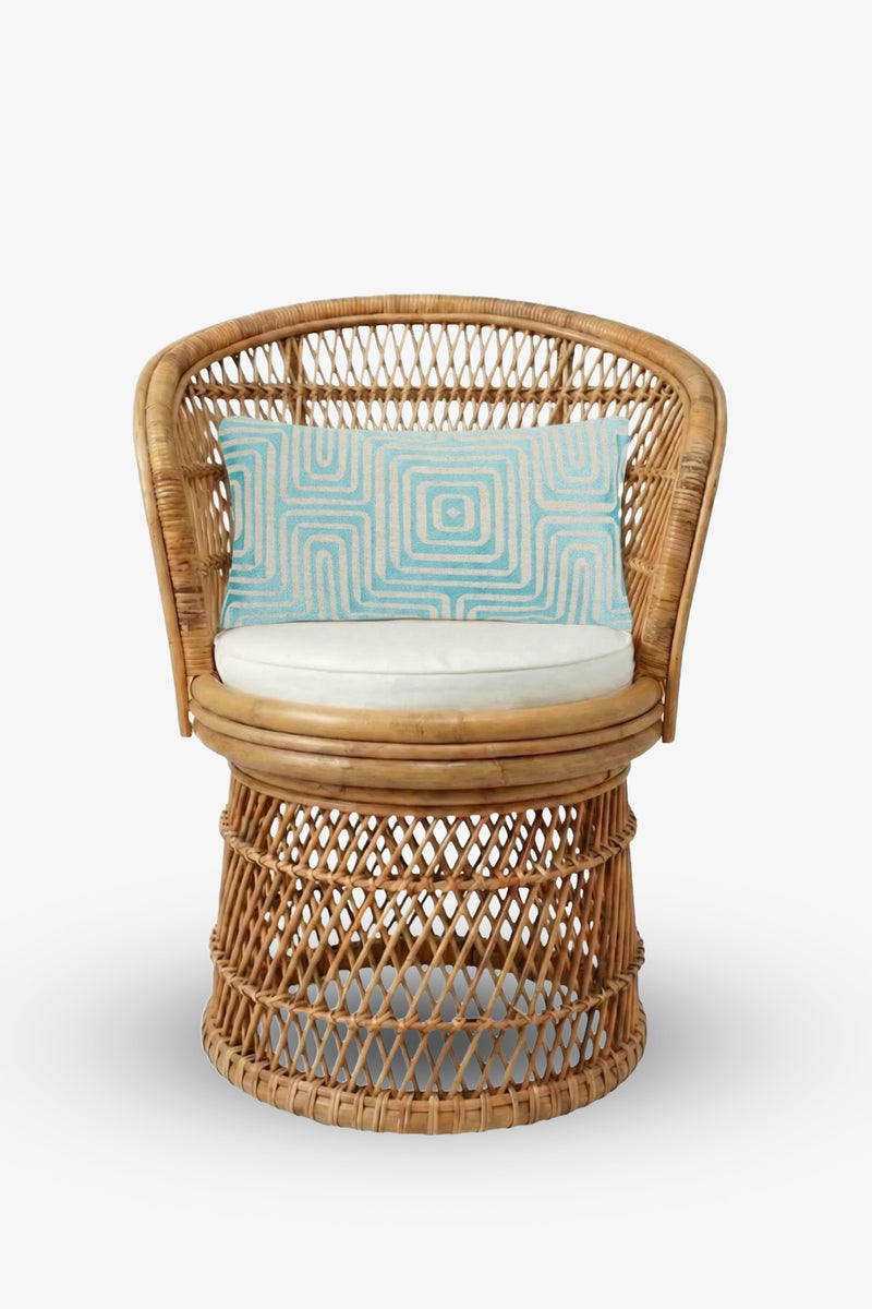 A round rattan chair with an open weave design sits on a plain white background, featuring a circular white cushion and the Amazing Maze Embroidered Lumbar Pillow—a rectangular light blue pillow adorned with a geometric swirl pattern.