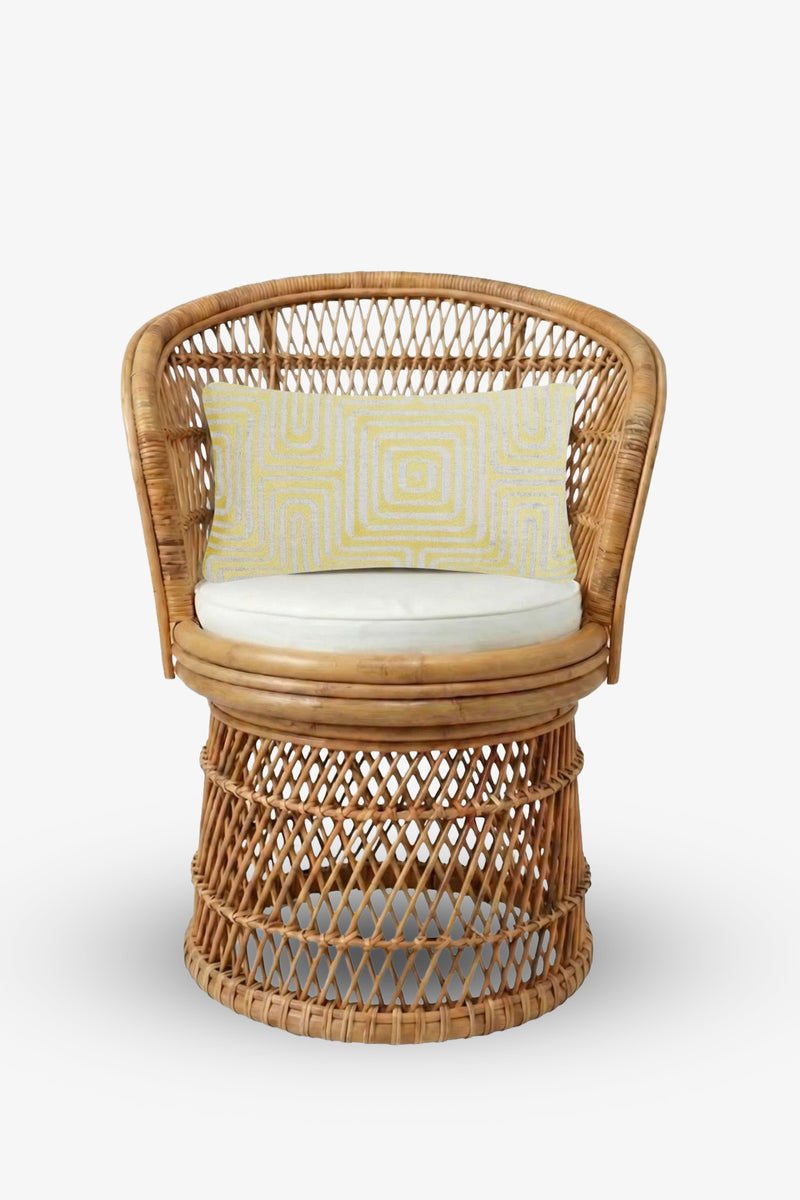 A wicker chair with a rounded back and open weave design is shown on a white background, featuring a white seat cushion and the Amazing Maze Embroidered Lumbar Pillow—a rectangular pillow adorned with a yellow geometric pattern.