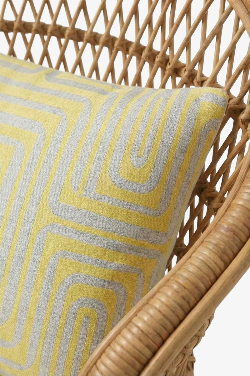 A close-up of a rattan chair featuring the Amazing Maze Embroidered Lumbar Pillow, showcasing its yellow and gray geometric design.