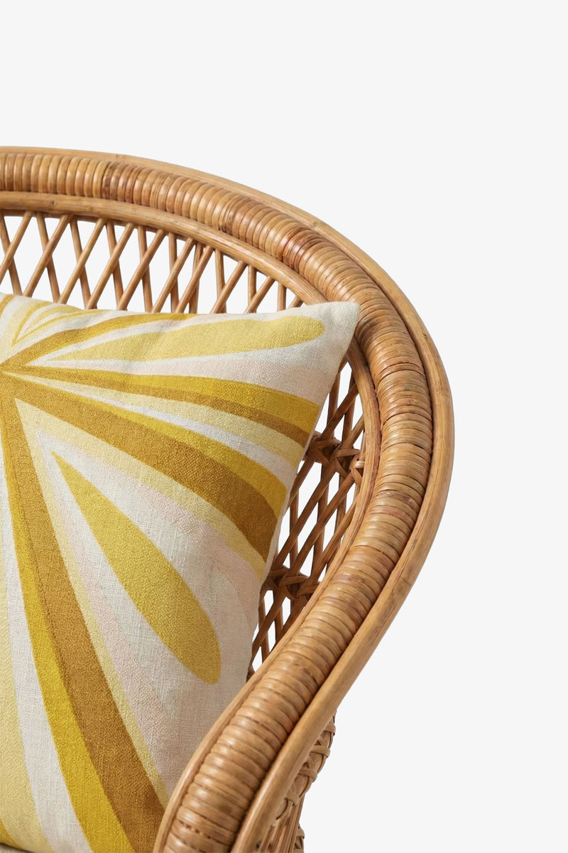 A close-up of a wicker chair with curved armrest and backrest, featuring the Shante Embroidered Square Pillow adorned with a yellow and white abstract sunburst pattern.