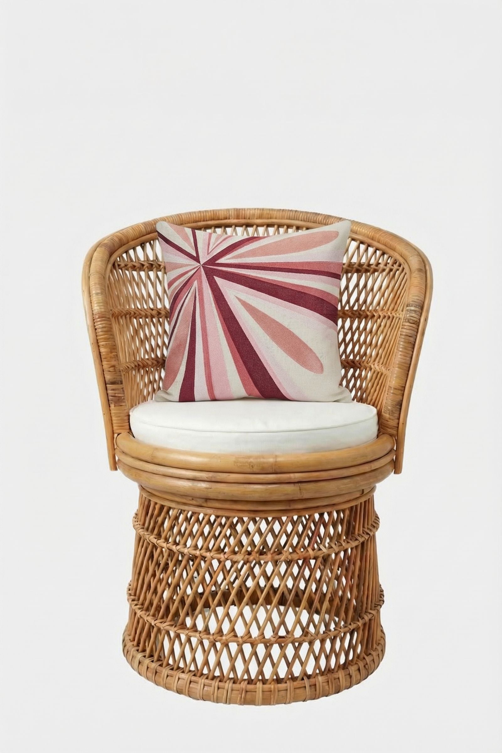 A round wicker chair with a curved back and white seat cushion features the Shante Embroidered Square Pillow, which displays a pink and red geometric sunburst pattern. The background is plain white.