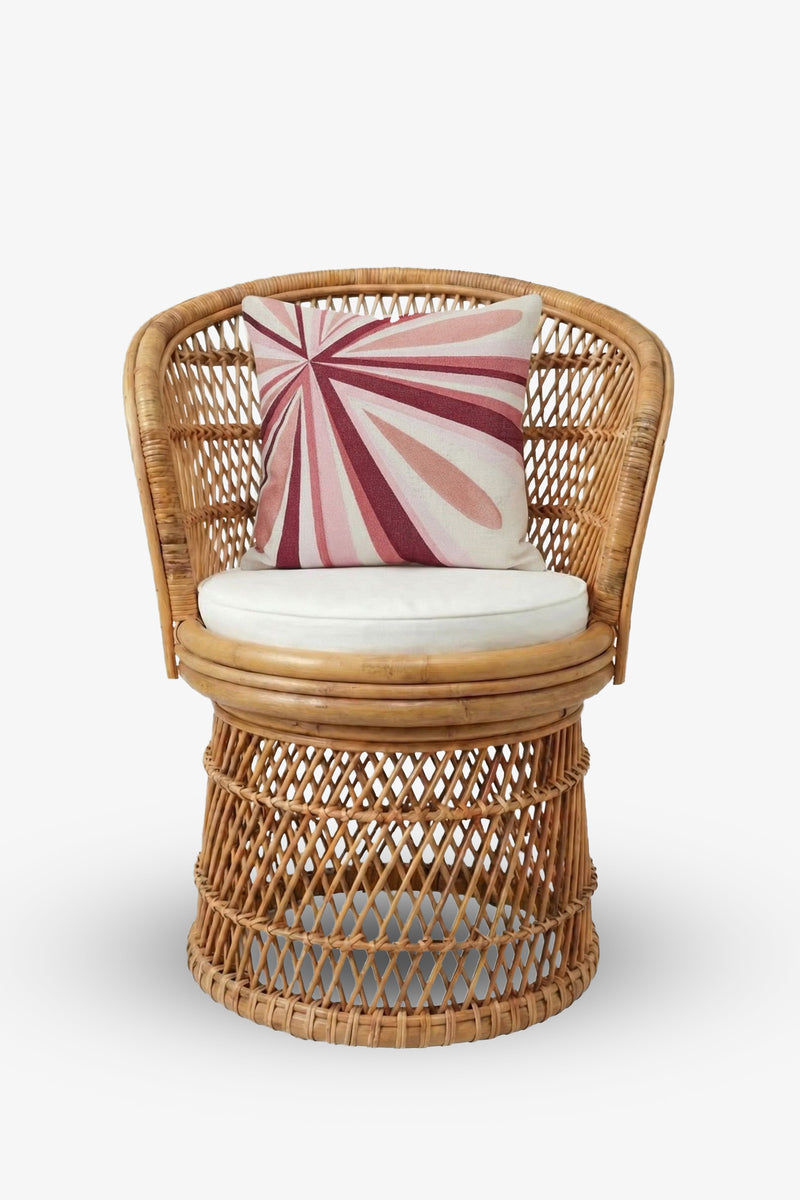 A round wicker chair with a white cushion and the Shante Embroidered Square Pillow, which features pink and maroon geometric sunburst patterns, set against a plain white background.