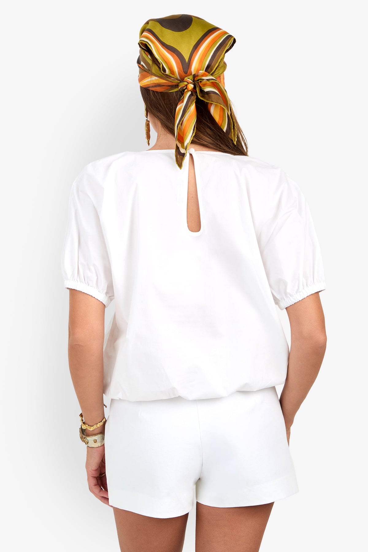Woman facing away, wearing the San Siro Top in white with matching shorts. She accessorizes with a vibrant patterned scarf and bracelets on her wrist.