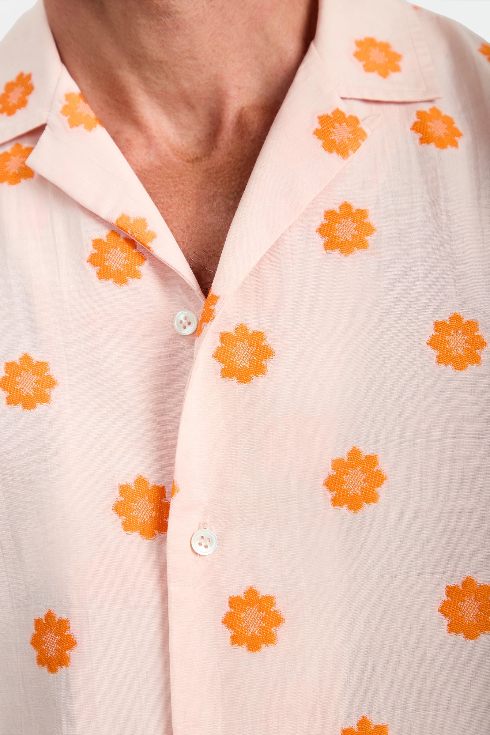 A close-up of a person wearing the Felix Camp Shirt - Daisy Dots, a pale pink, semi-sheer shirt with an open collar, partial buttons, and orange embroidered daisy dot patterns.