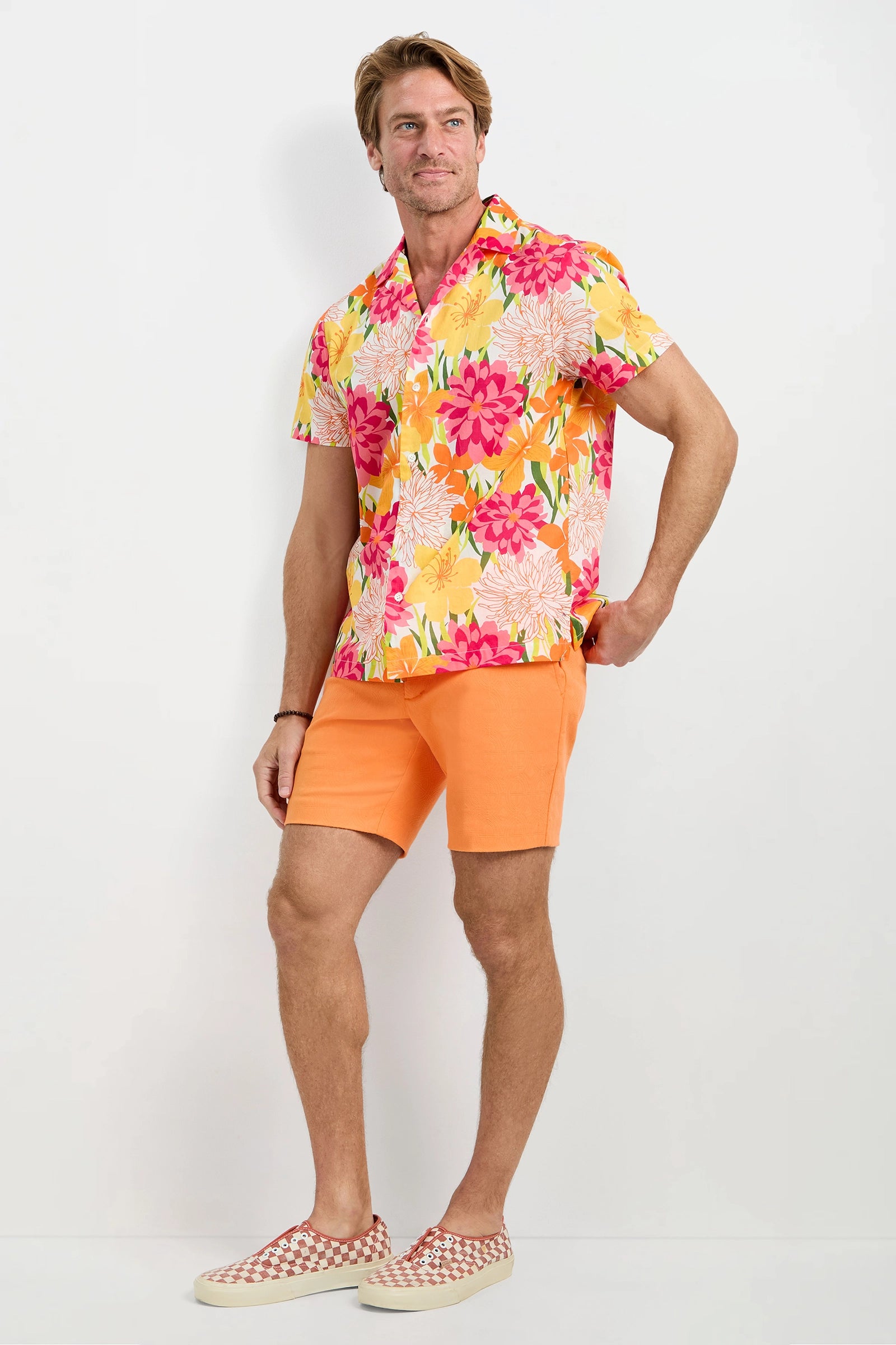 A man stands against a white background wearing the Felix Camp Shirt - Como, featuring a yellow, pink, and white floral print, paired with orange shorts and red-and-white checked slip-on shoes.