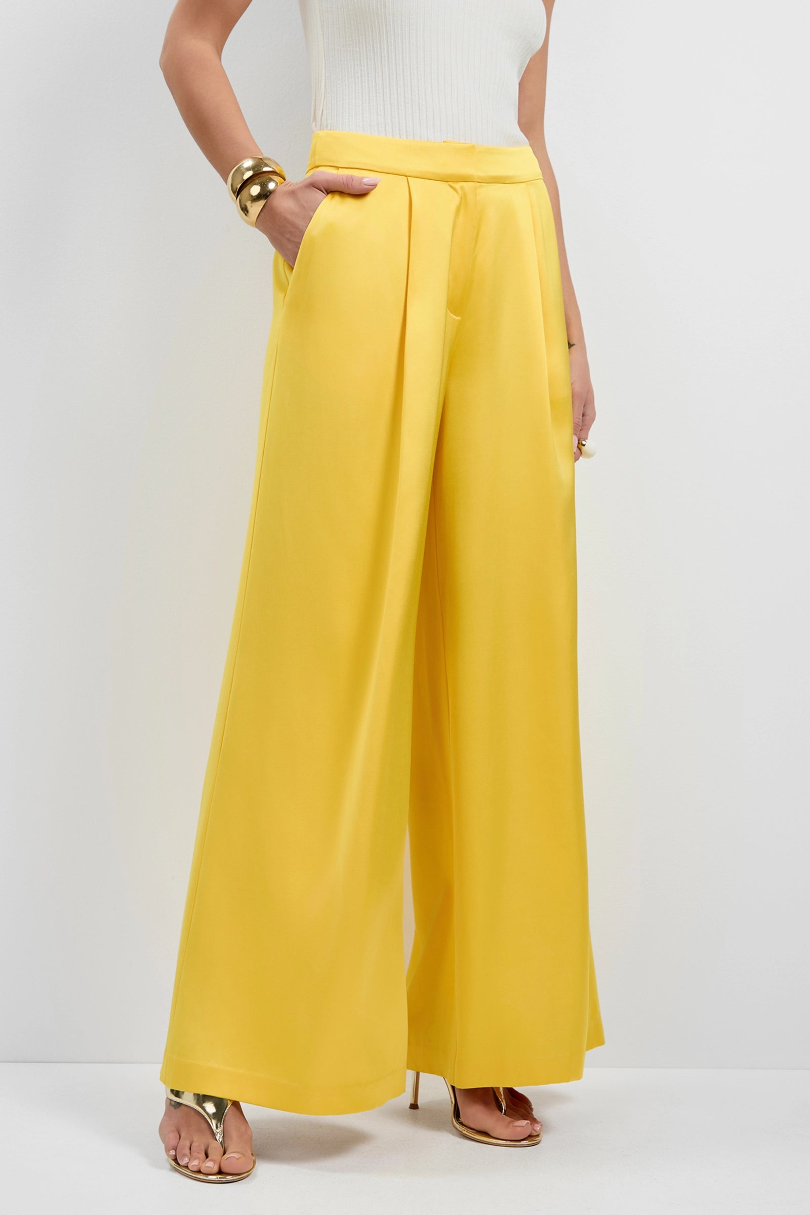 A person wearing Pontaccio Satin Pants—high-waisted, wide-leg yellow trousers—with a white sleeveless top, gold bracelets, and gold high-heeled sandals, standing against a plain white background.