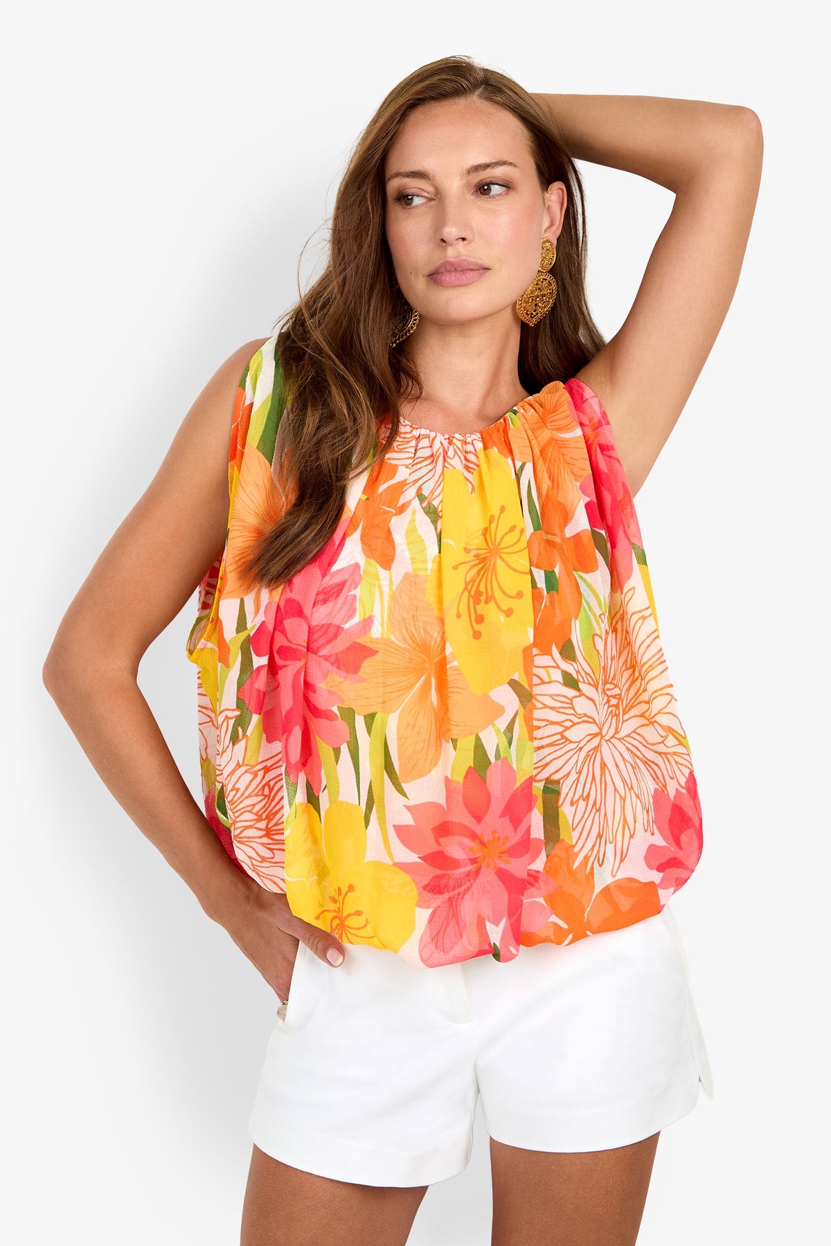 Model wears the Brera Top (Wholesale), a sleeveless, colorful floral top, paired with white shorts. She stands confidently against a plain background with one hand on her hip and the other arm raised.