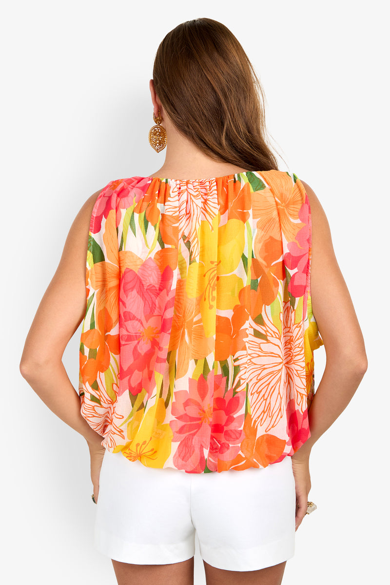 Woman facing away, wearing the Brera Top (Wholesale)—a colorful, sleeveless, floral blouse in orange, pink, and yellow—paired with white shorts. She has brown hair down and large round gold earrings.