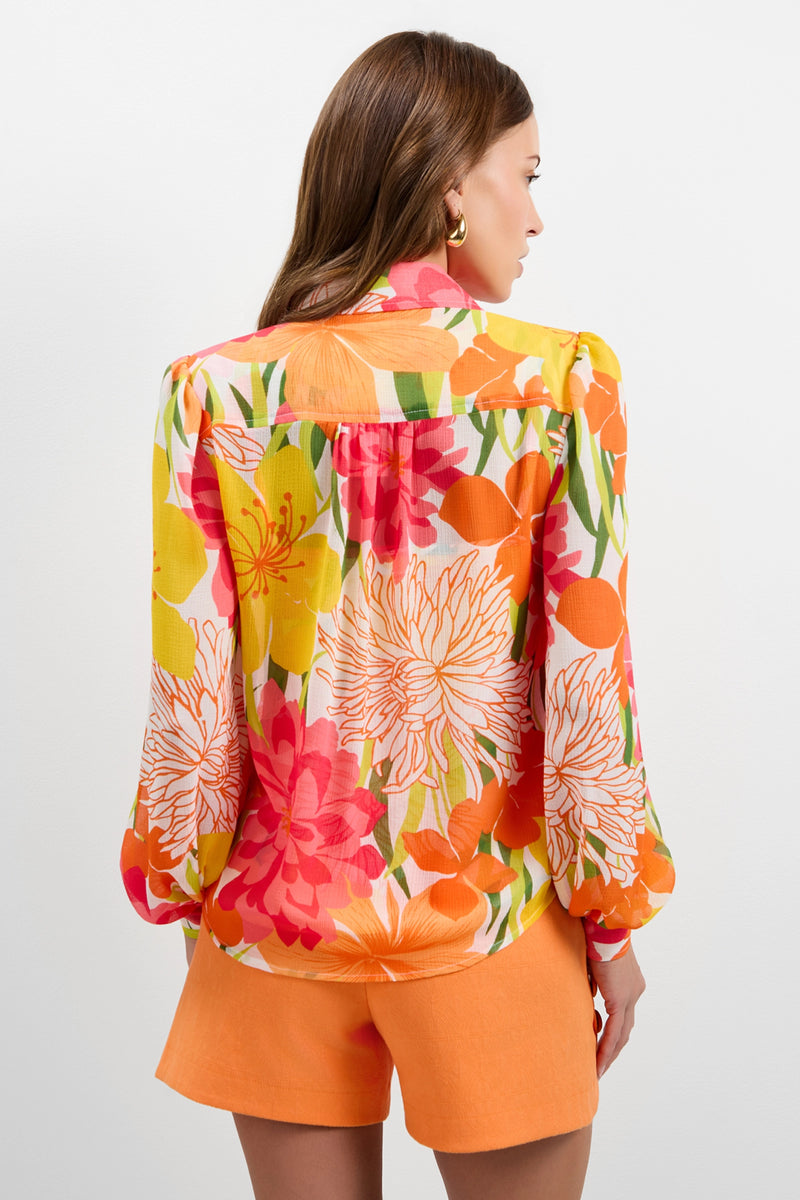 A woman with long brown hair faces away, showcasing the back of the Monte Balloon Sleeve Top—a colorful floral blouse with orange, yellow, and pink flowers—paired with orange shorts against a plain white background.