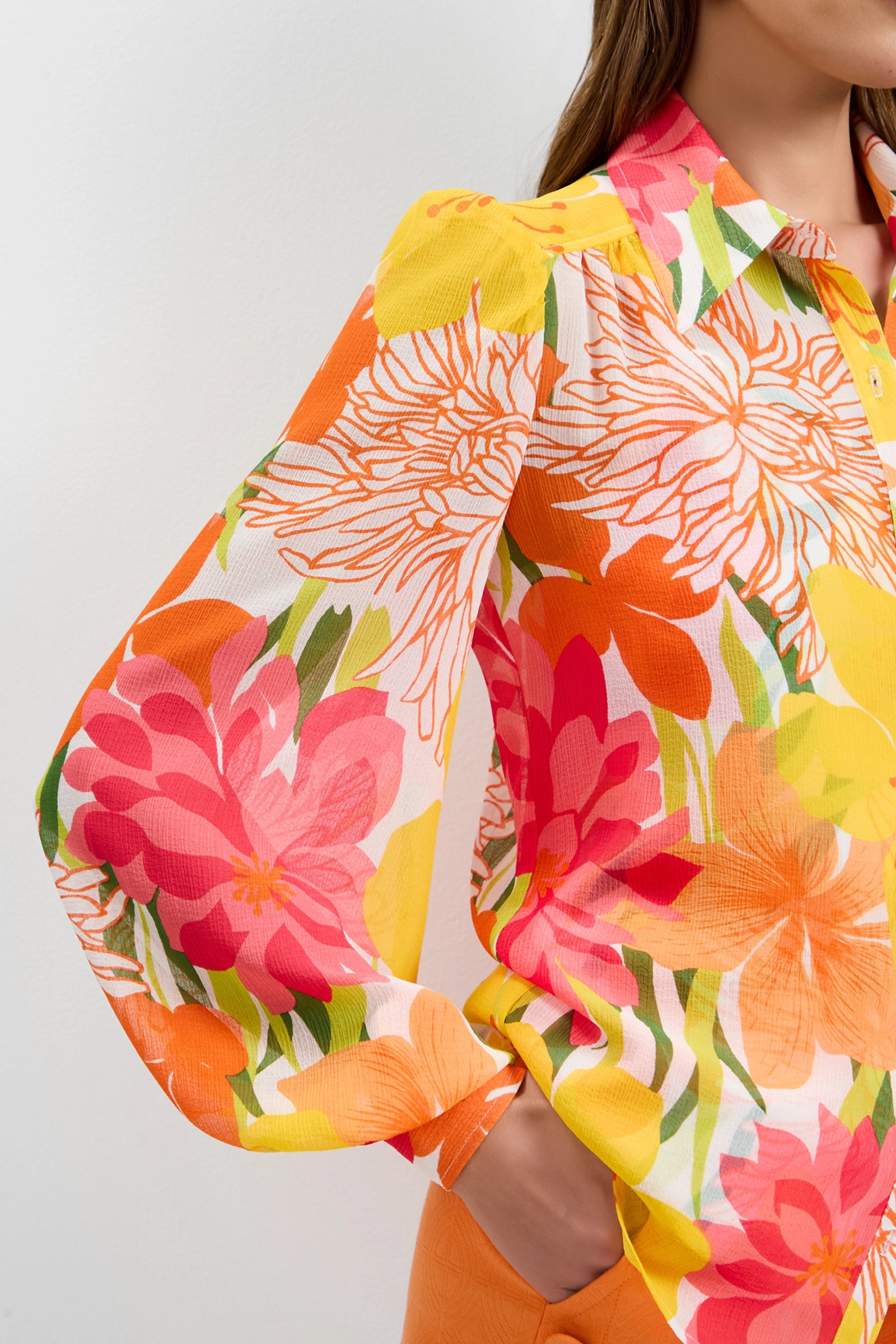 A woman wears the Monte Balloon Sleeve Top, a long-sleeved, button-up blouse with a vibrant floral print in orange, yellow, pink, and green, paired with orange pants. Her right hand is in her pocket and her head is partially out of frame.