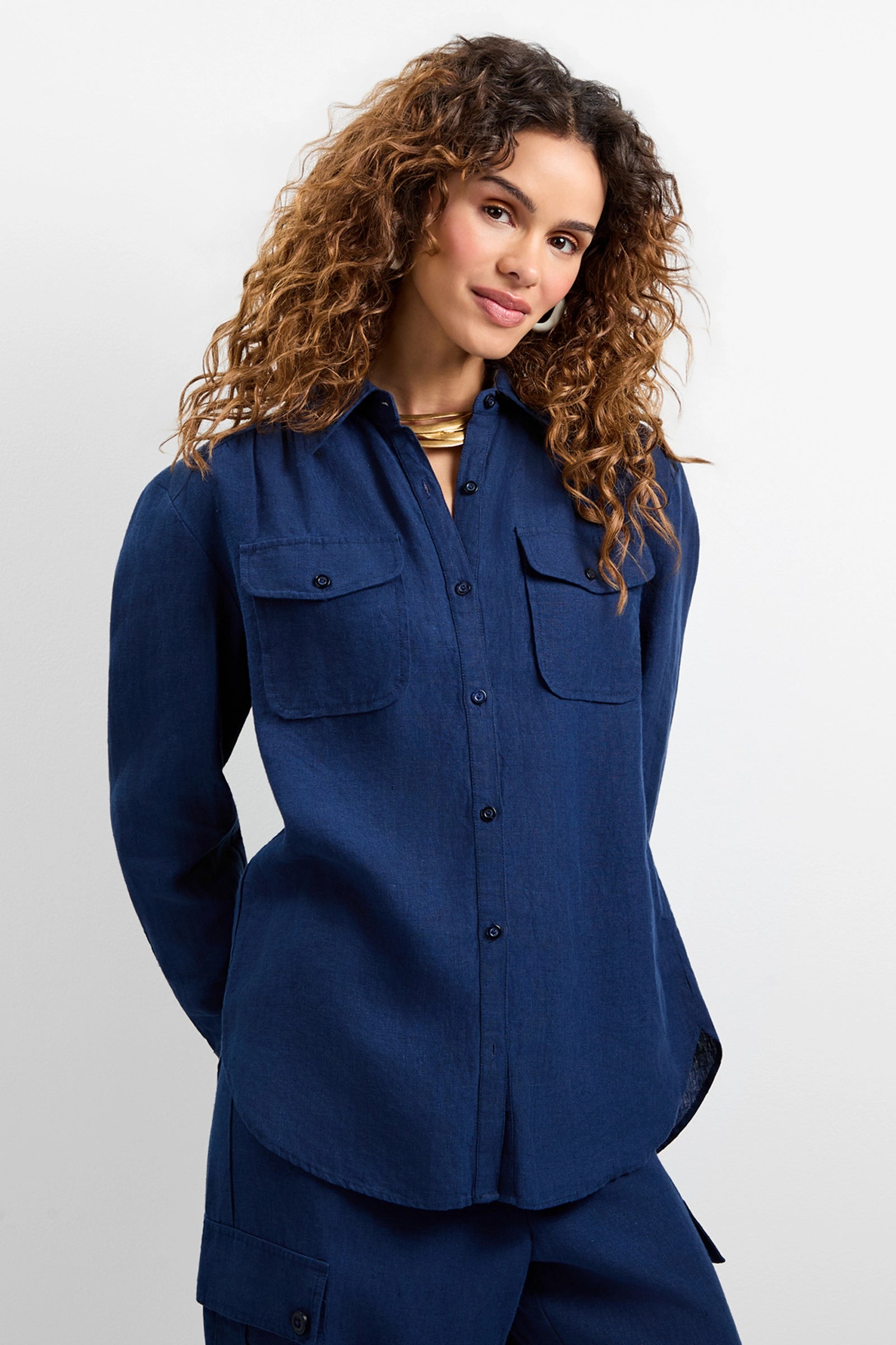 A woman with curly brown hair, smiling slightly, wears the Barona Top—a dark blue button-up shirt with front pockets—and matching pants while standing against a light background.