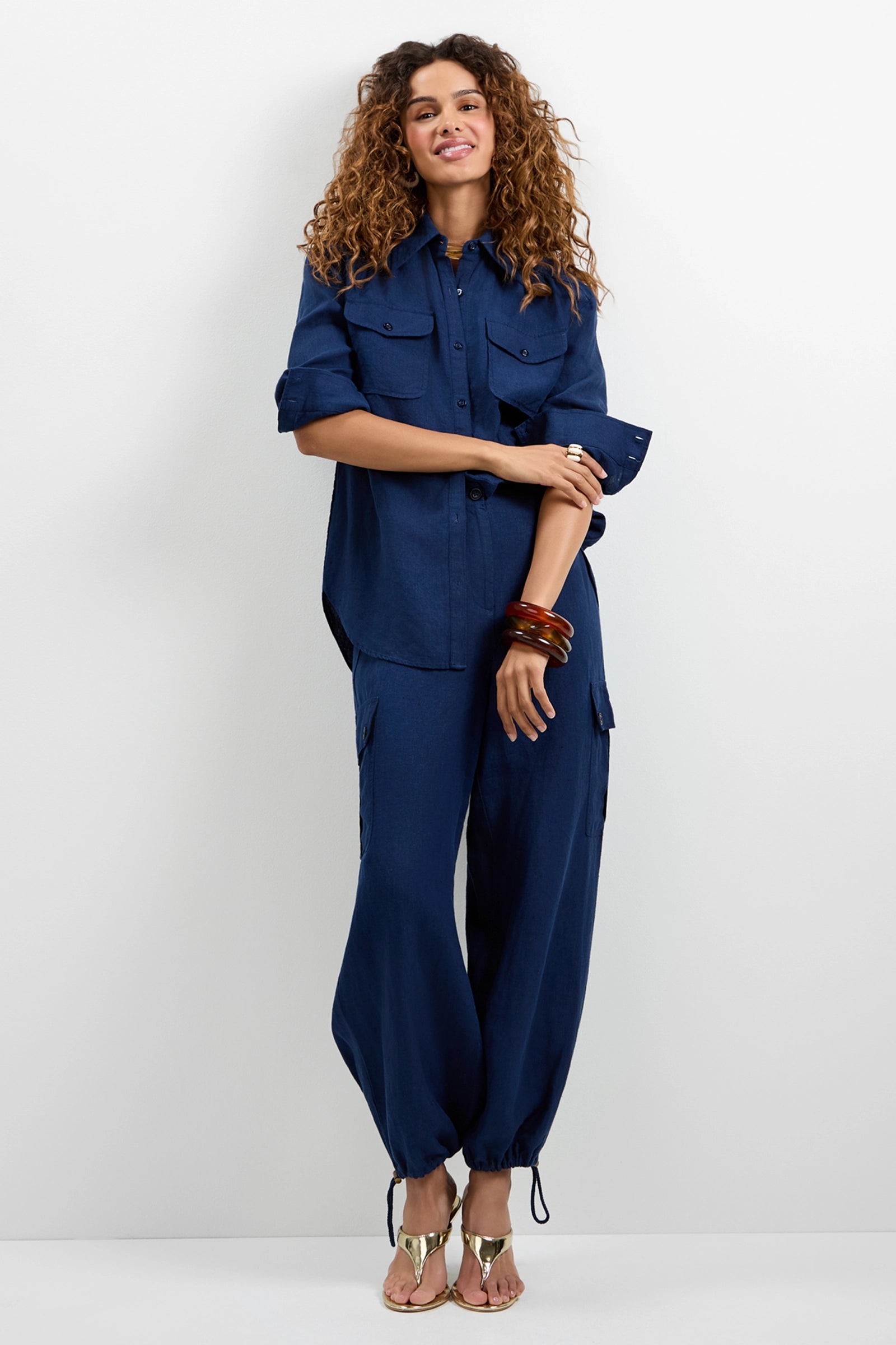 A person with curly hair wears the Barona Top, a loose-fitting navy blue button-up shirt, and matching jogger pants with drawstring hems, styled with gold sandals and stacked bangles against a plain white background.