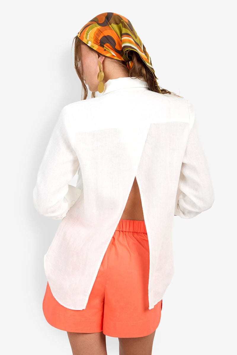 A woman wears a patterned headscarf, gold earrings, and the Barona Top (Wholesale) with a back slit, paired with bright orange shorts, shown from behind against a plain background.