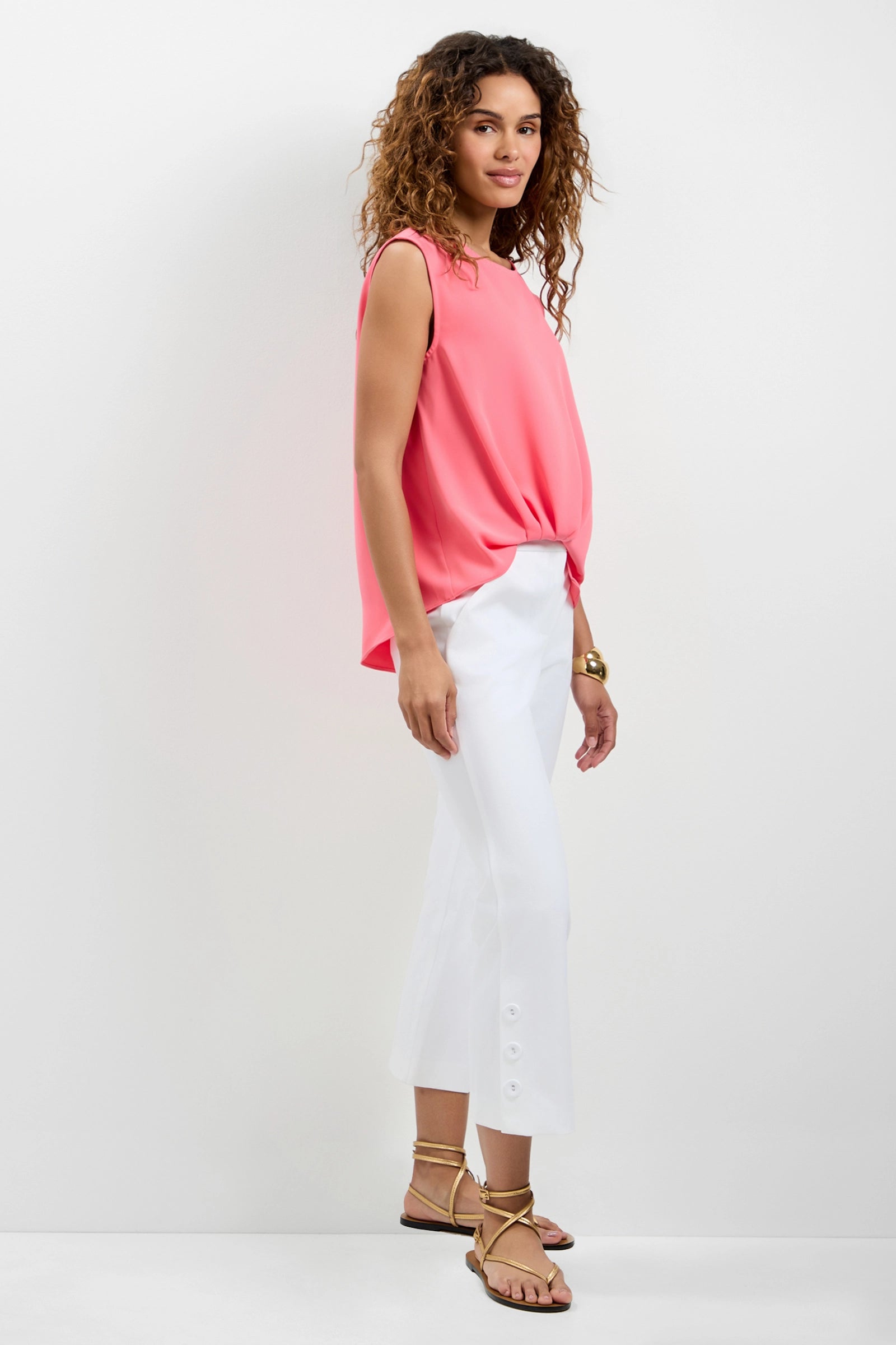 A woman with curly hair poses against a white background, wearing the Aprica Boatneck Top in pink, white cropped pants with button details at the hem, a gold bracelet, and strappy flat sandals.