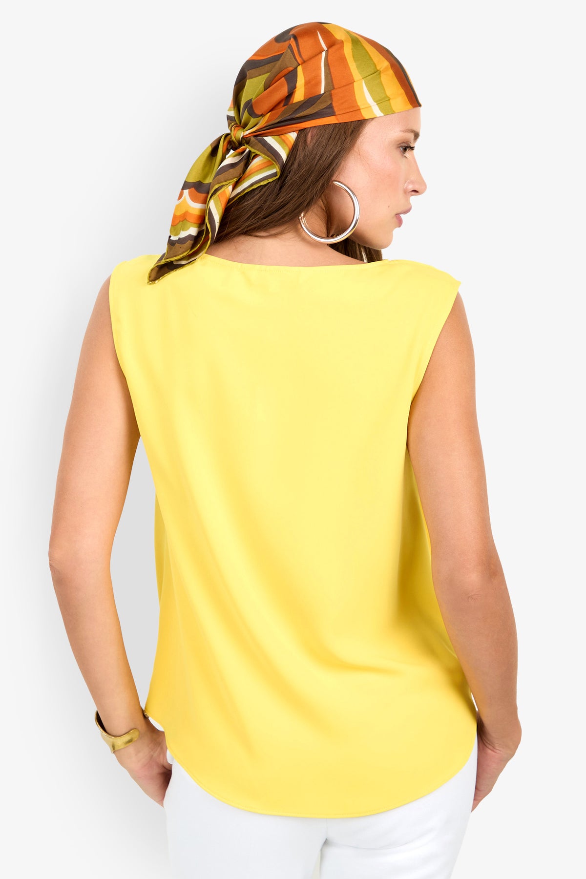 A woman faces away wearing the Aprica Boatneck Top (Wholesale) in yellow, white pants, large hoop earrings, and a vibrant orange-green-yellow headscarf. Her right hand is in her pocket against a plain white background.