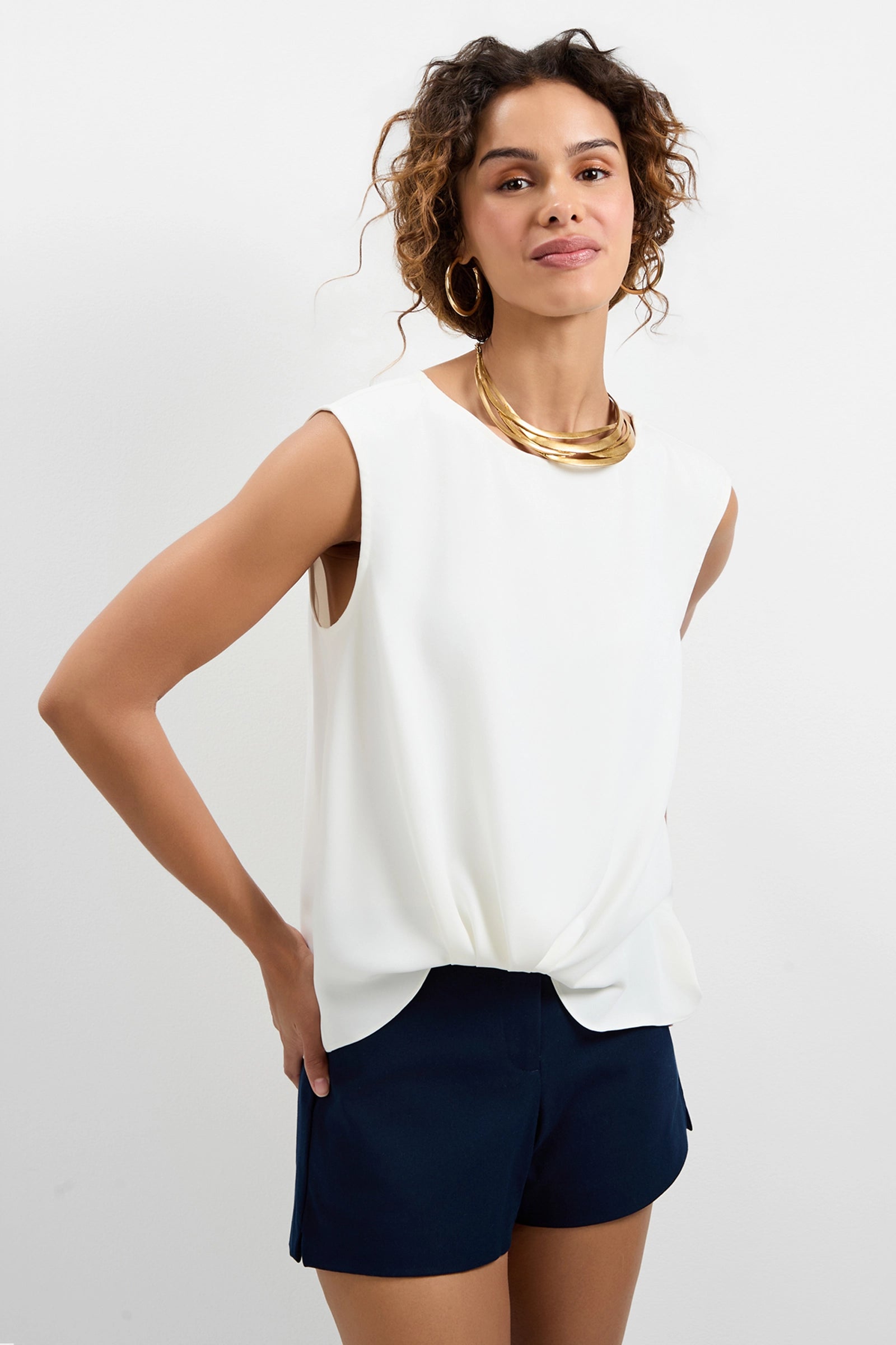 A woman with curly hair and a confident look stands against a plain white background, wearing the Aprica Boatneck Top, dark navy shorts, large hoop earrings, and a gold statement necklace.